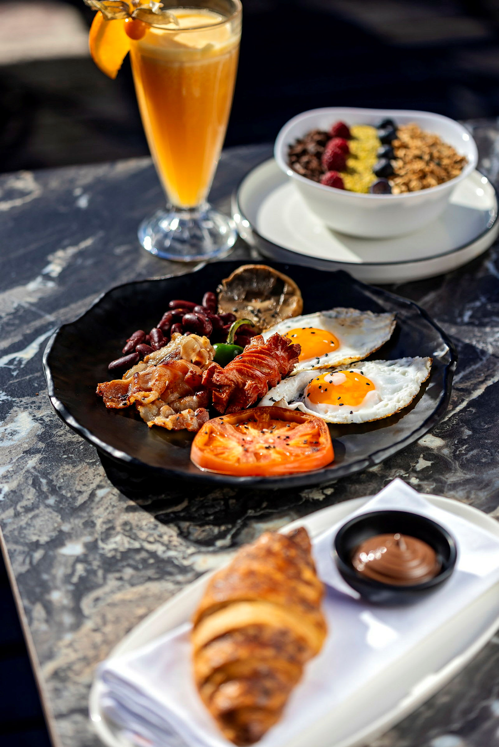 En tallerken med morgenmad bestående af bacon, æg, tomat og grøntsager. En croissant med chokoladecreme samt en skål med morgenmadsprodukter og et glas juice er også til stede. (AI-genereret tekst)