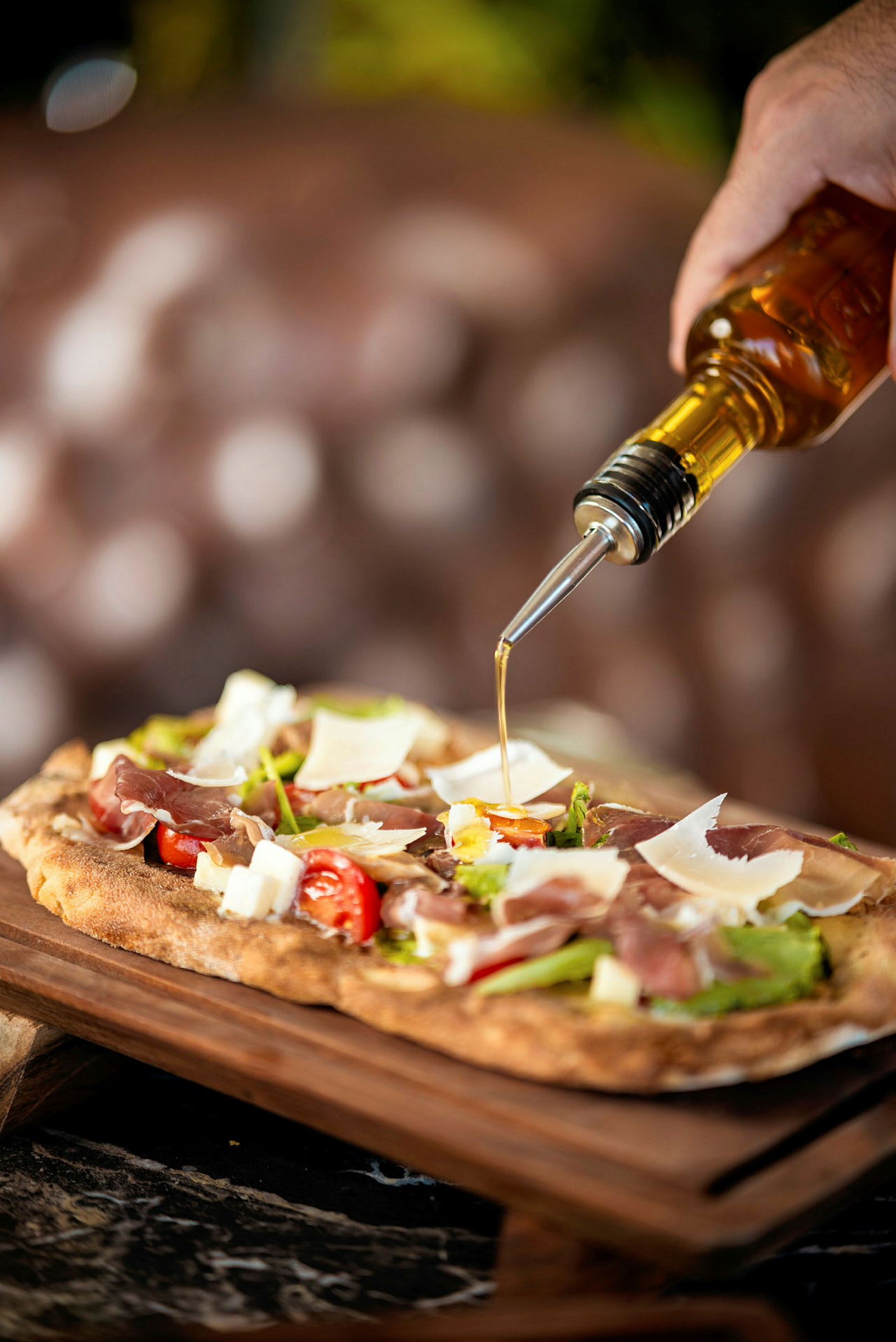 En hånd hælder olivenolie fra en flaske over en pizza med skinke, tomater og parmesanost. (AI-genereret tekst)