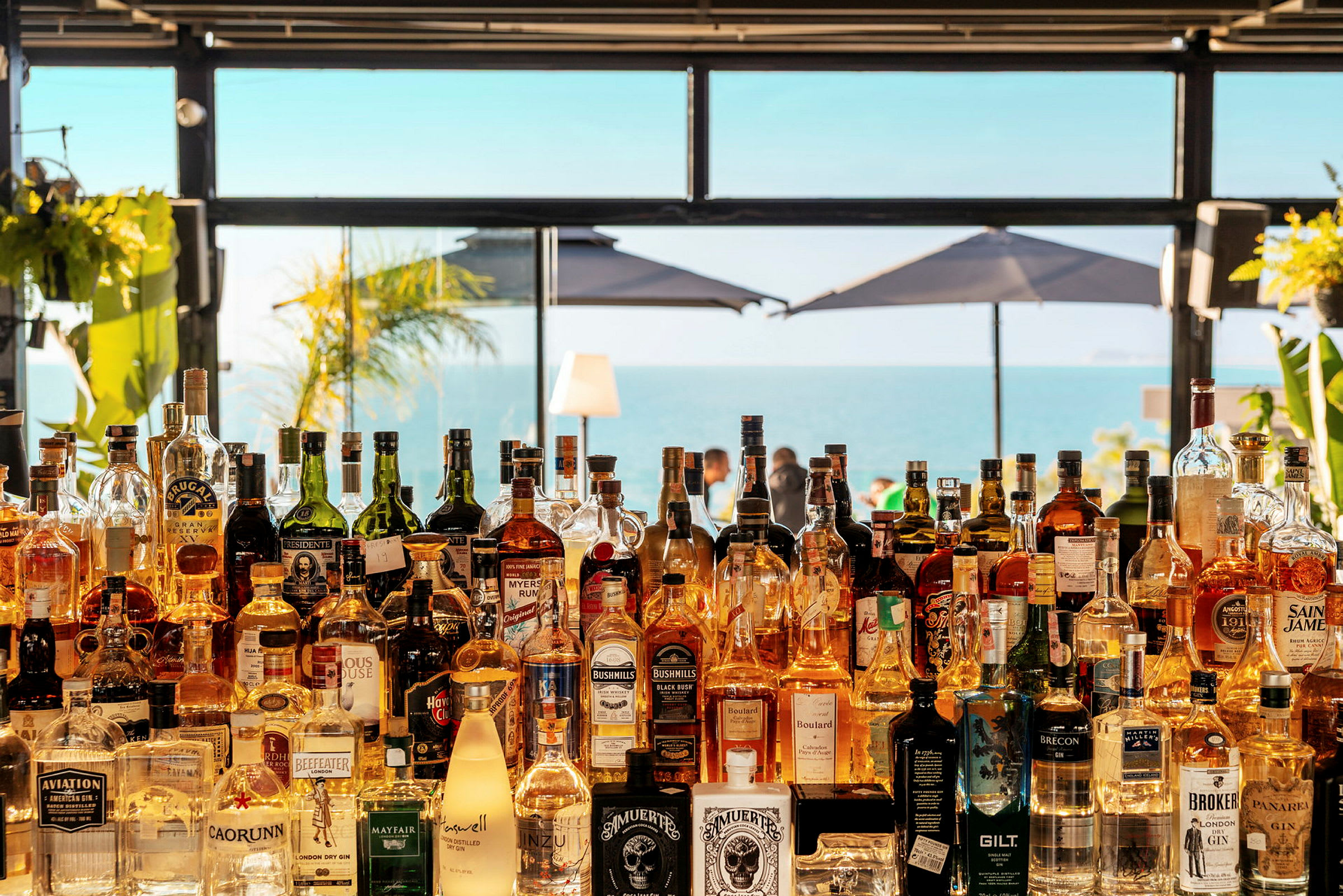 Udsigt over en strandbar med flasker på en hylde og havet i baggrunden. (AI-genereret tekst)