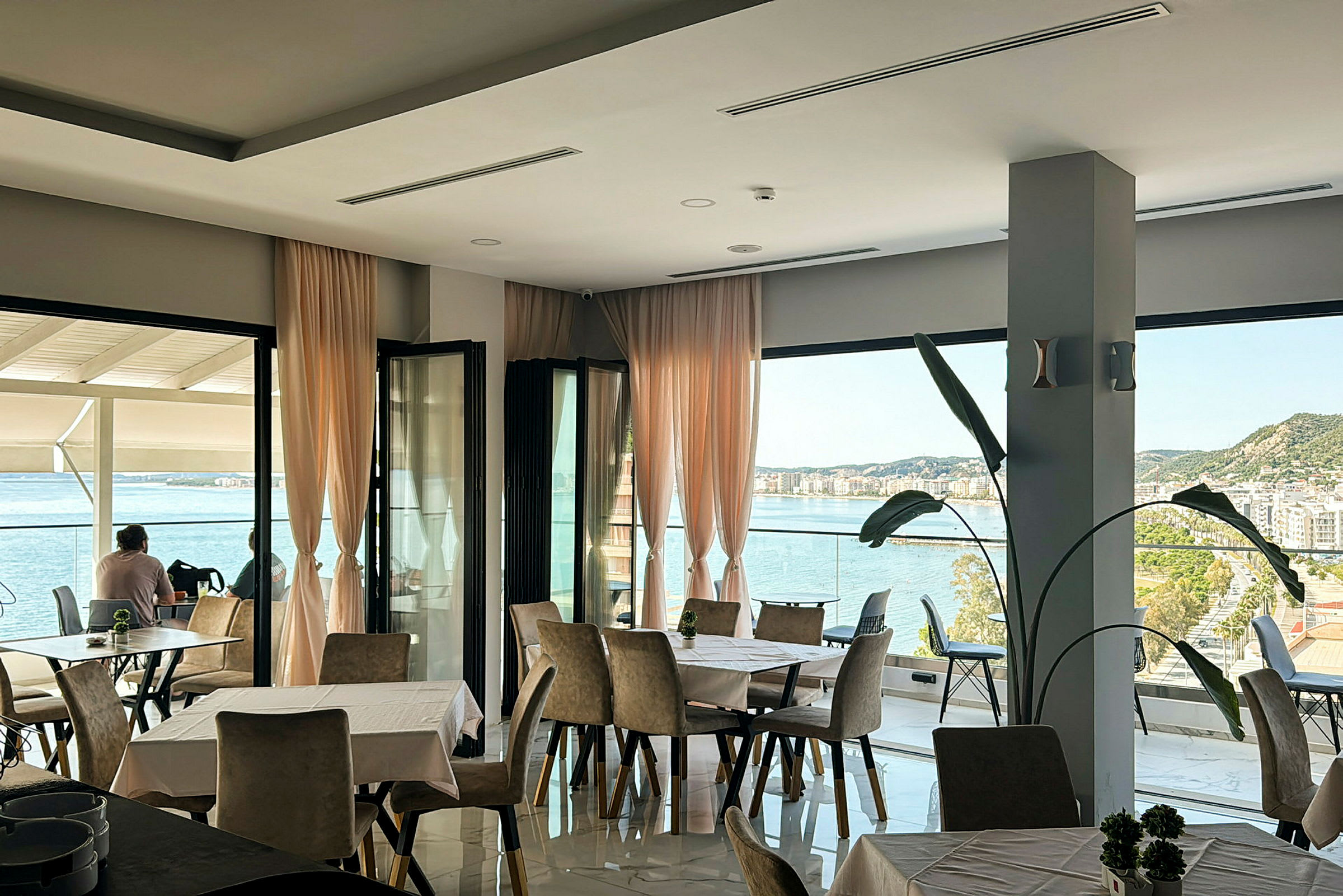 Et restaurantområde med store vinduer og udsigt over havet og en by. (AI-genereret tekst)