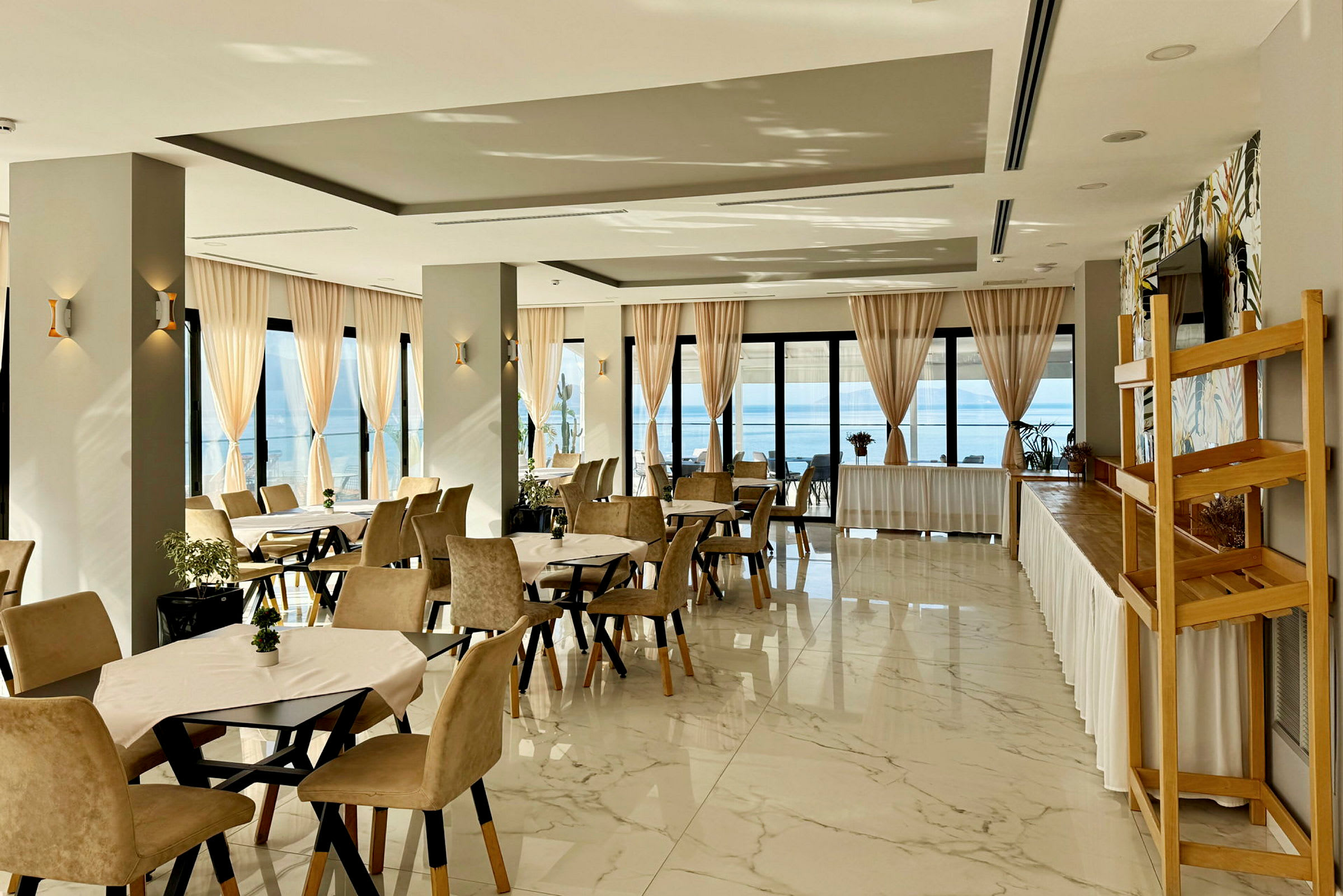 Restaurant med dækkede borde og beige stole, store vinduer med lyse gardiner og havudsigt. (AI-genereret tekst)