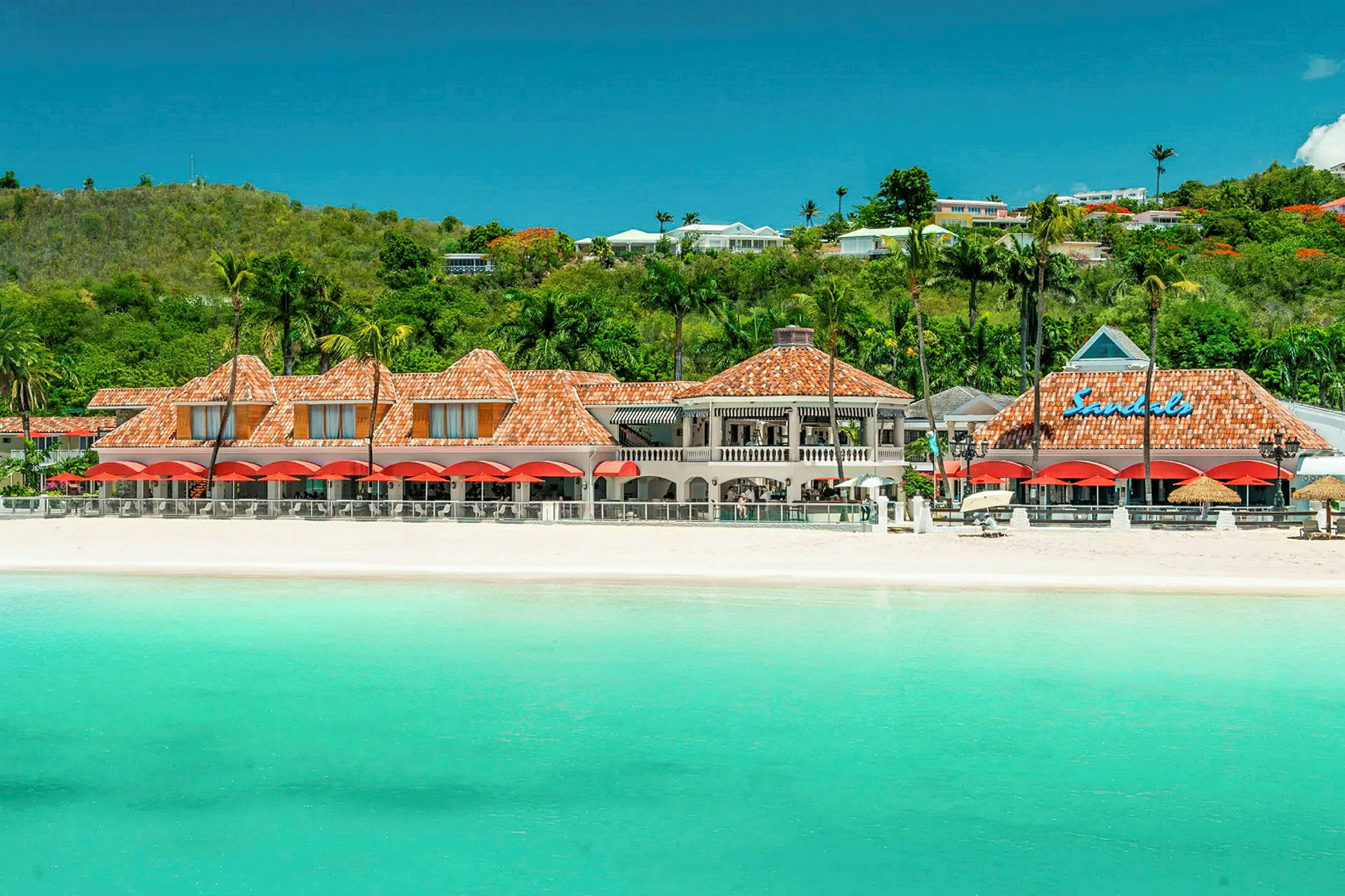 En strand med turkis vand foran en resortbygning med røde tage og grønt i baggrunden. (AI-genereret tekst)