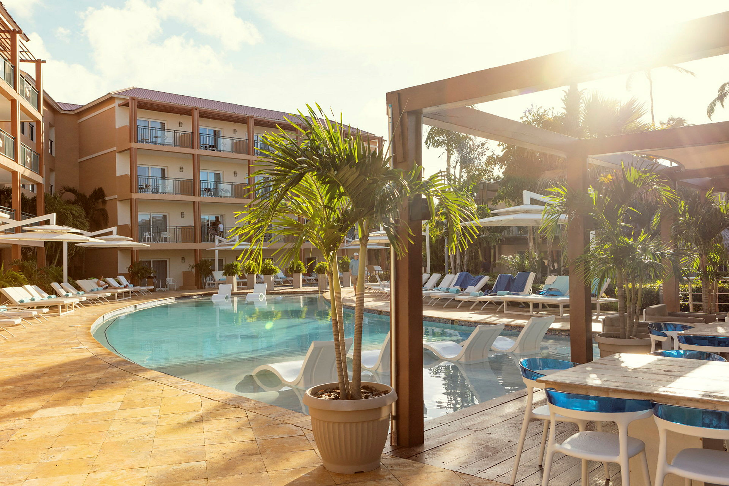 Billede af et hotelområde med en pool omgivet af liggestole og parasoller. Foran ses borde og stole placeret ved en træterrasse. (AI-genereret tekst)
