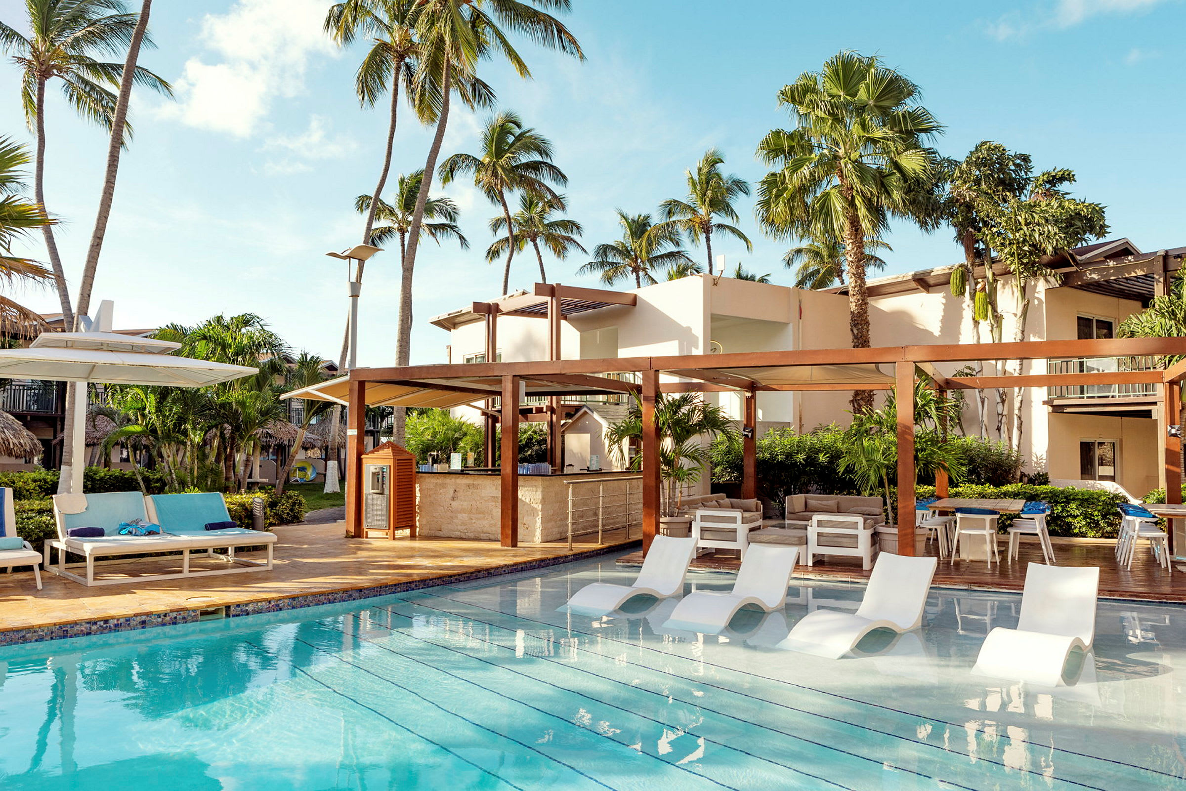 Billedet viser en udendørs pool ved siden af en moderne ferieresort omgivet af palmer. (AI-genereret tekst)