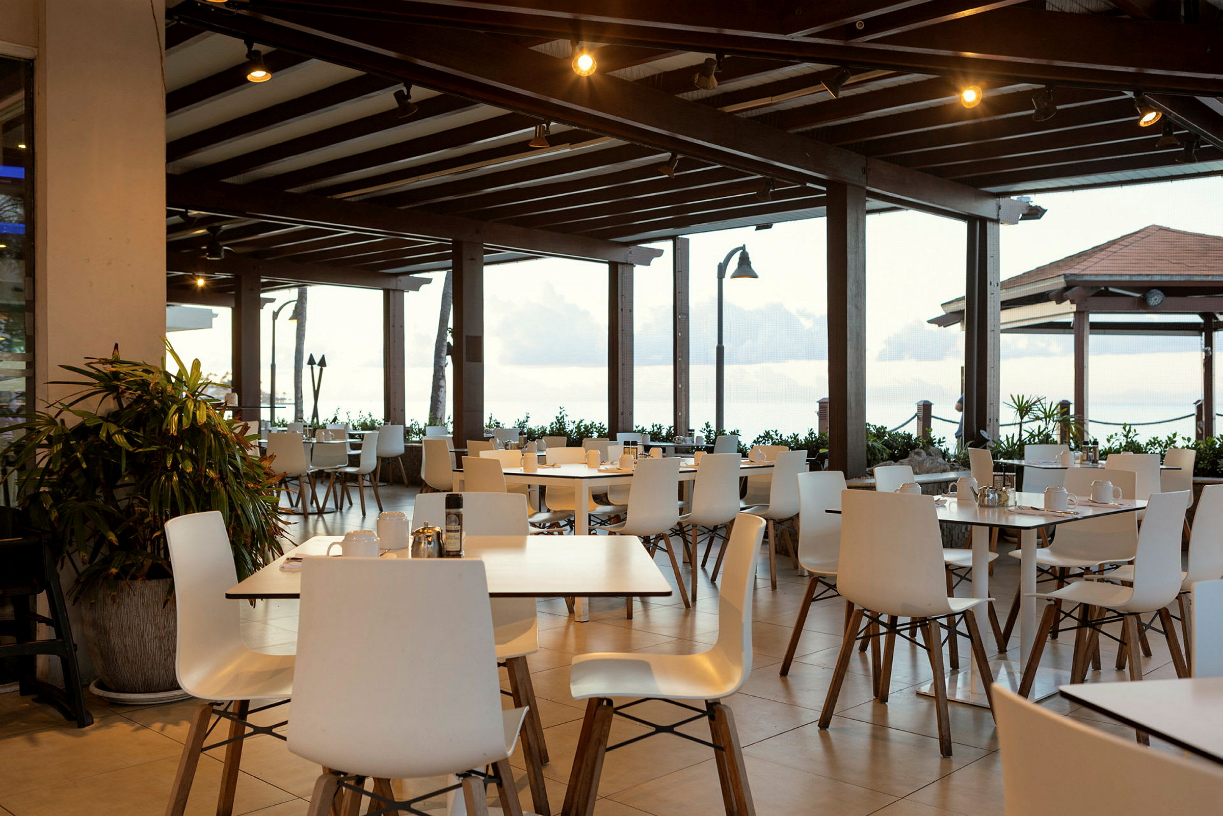 En restaurantterrasse med hvide stole og borde under et trætag. Udsigt over havet bagved. (AI-genereret tekst)
