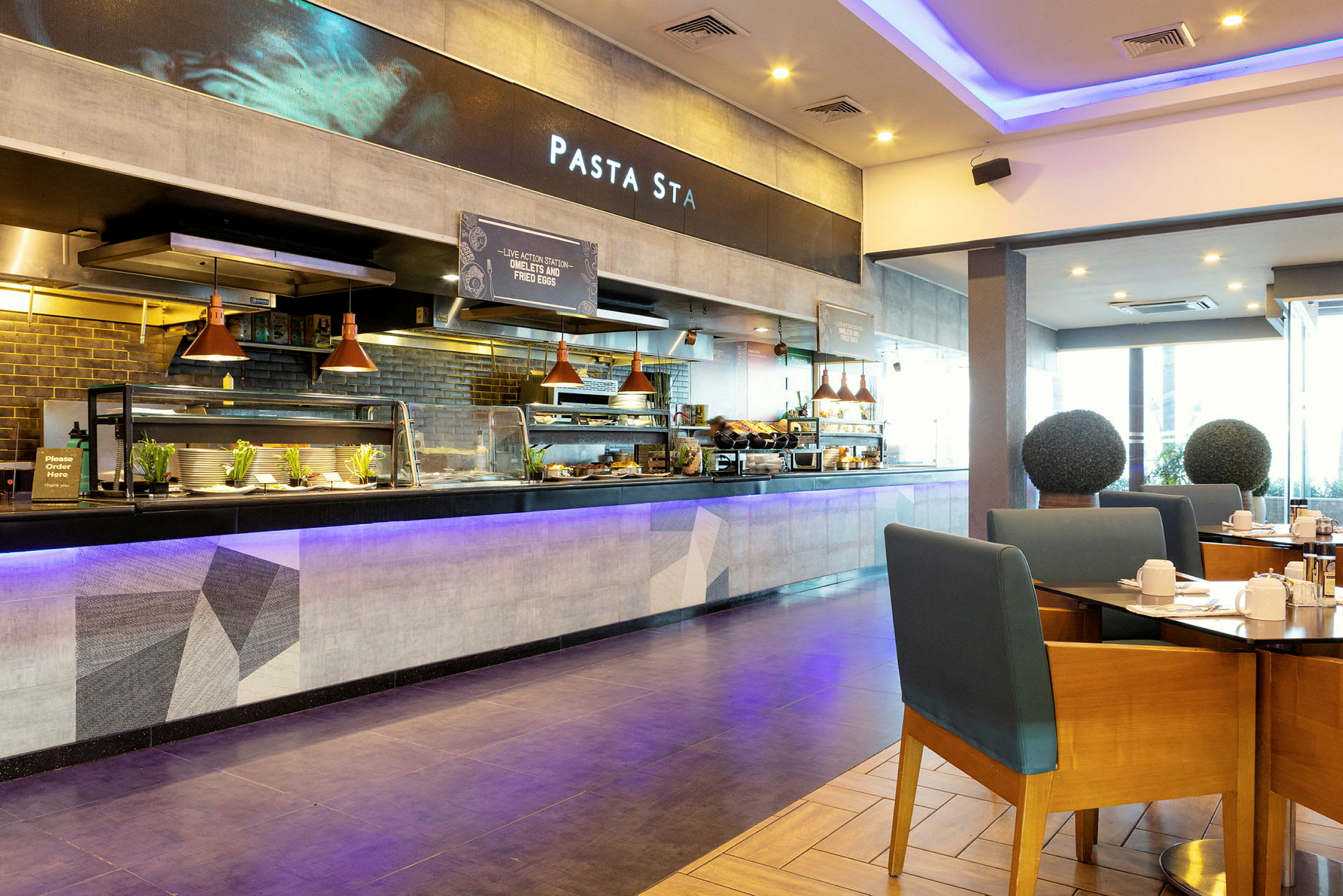 En restaurant med en åben serveringsdisk og siddepladser. Borde med stole ses i forgrunden, og bag serveringsdisken vises et skilt med teksten 'Pasta Sta'. (AI-genereret tekst)