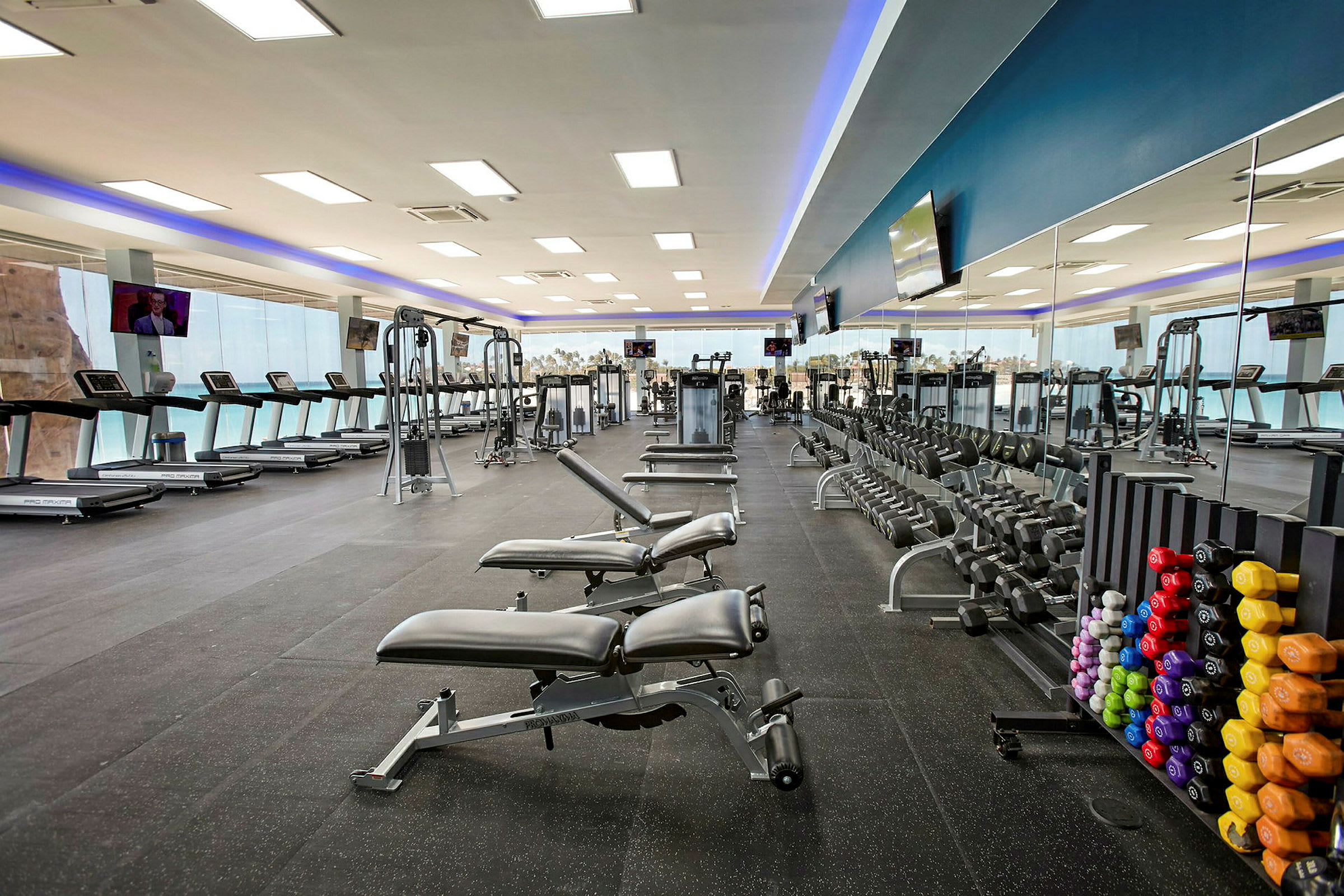 Et fitnesscenter med træningsudstyr som løbebånd, håndvægte og træningsmaskiner. En spejlvæg ses på den ene side. (AI-genereret tekst)