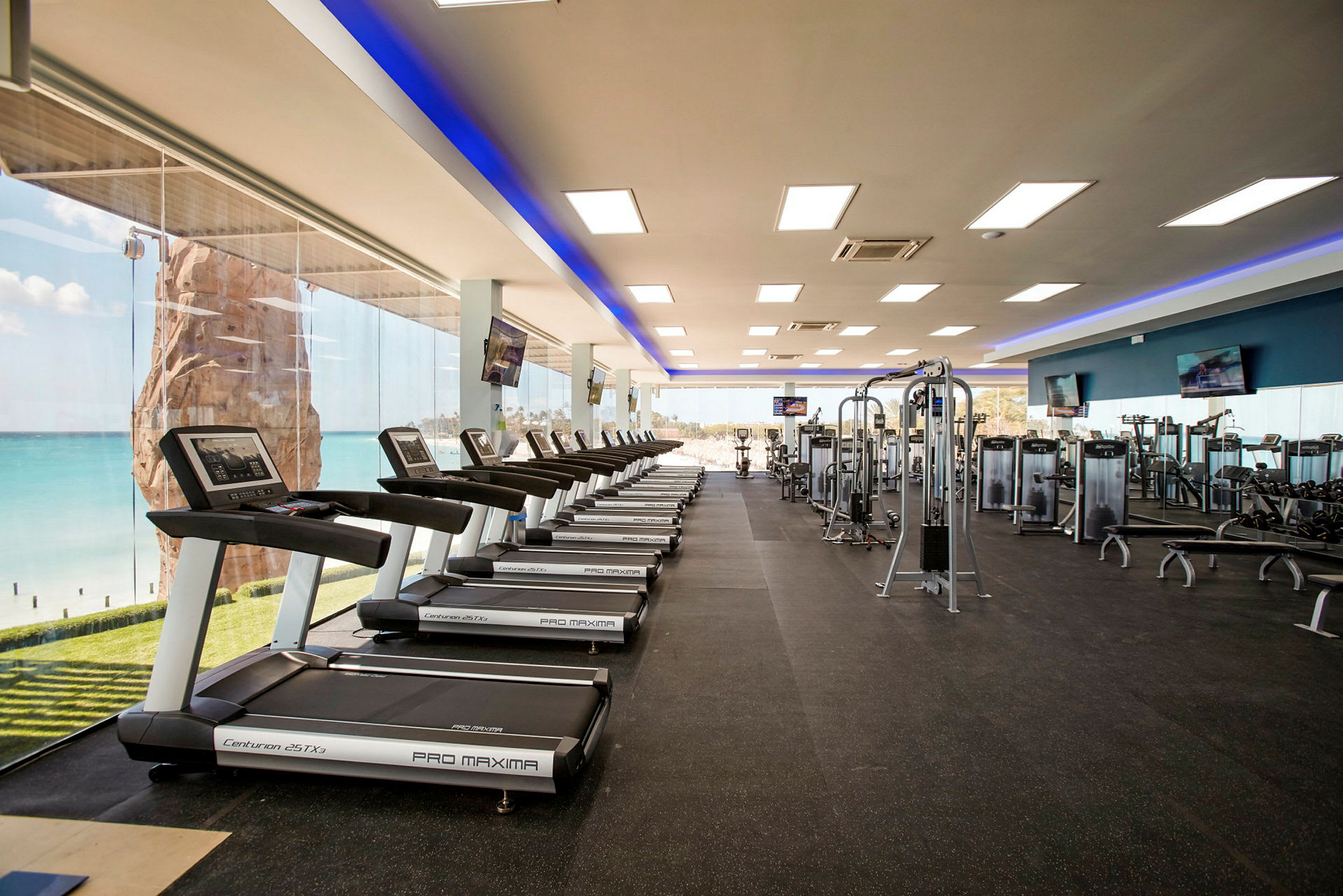 Fitnesscenter med løbebånd og andre træningsmaskiner, stor vinduesvæg med udsigt over havet. (AI-genereret tekst)