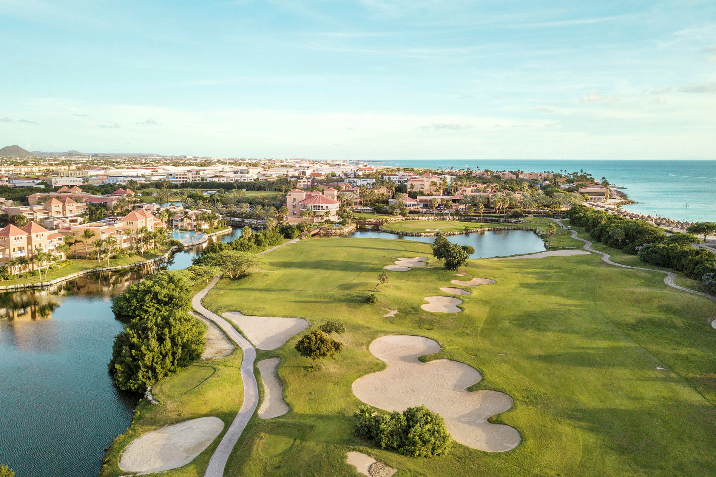 Golfbane med sandbunkere og grønne græsarealer nær vandløb og strand. Bebyggelse i baggrunden. (AI-genereret tekst)