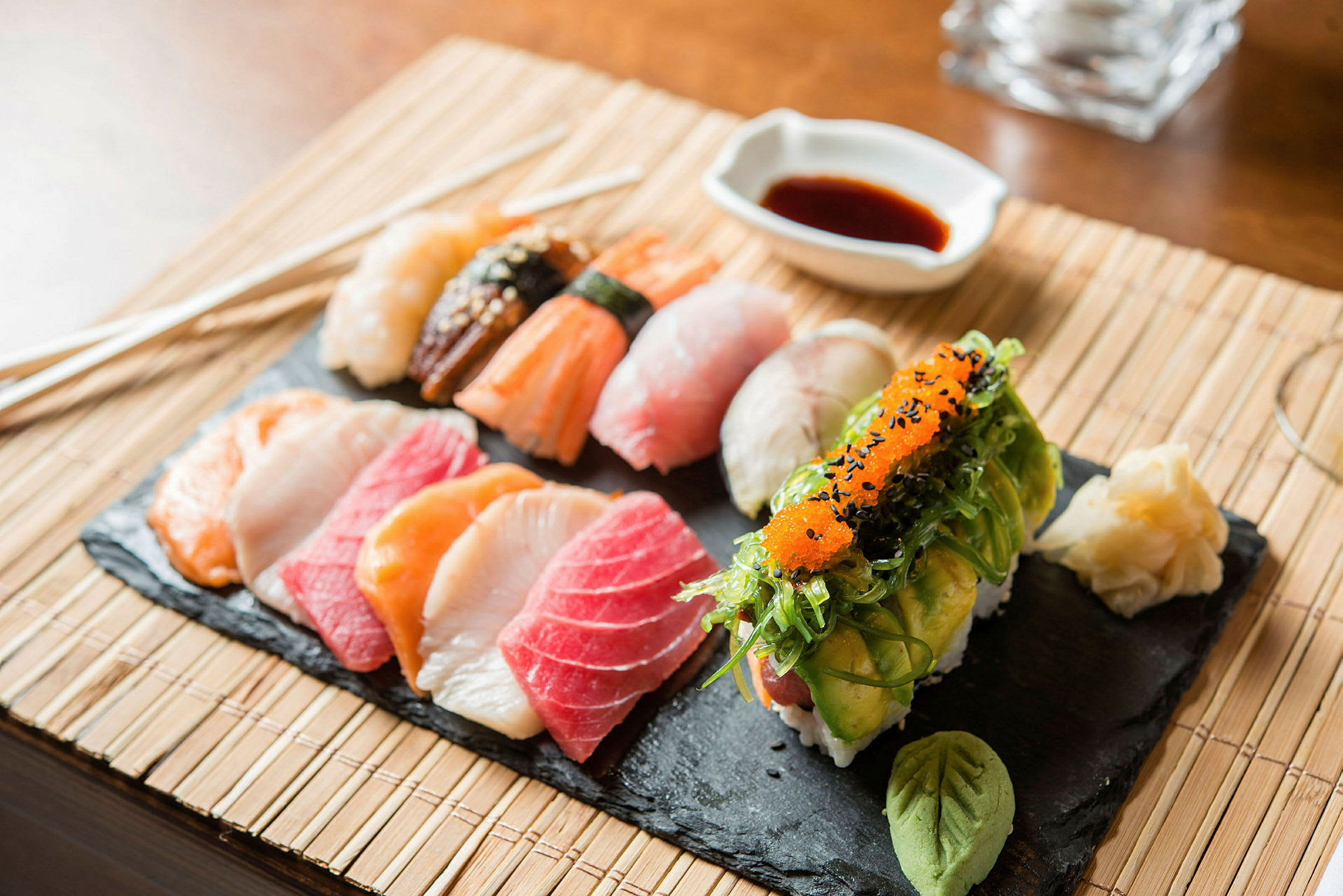 En skiferplade med forskellige slags sushi, wasabi og syltet ingefær, samt sojasovs i en skål. (AI-genereret tekst)