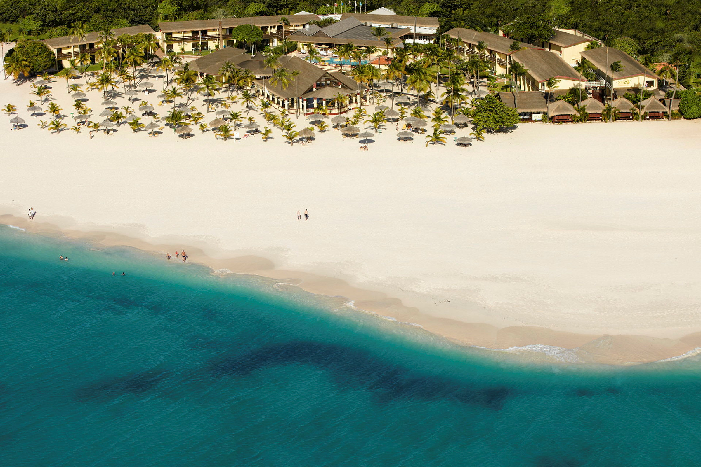 Luftfoto af en hvid sandstrand med blåt hav foran et hotelområde omgivet af palmer. (AI-genereret tekst)