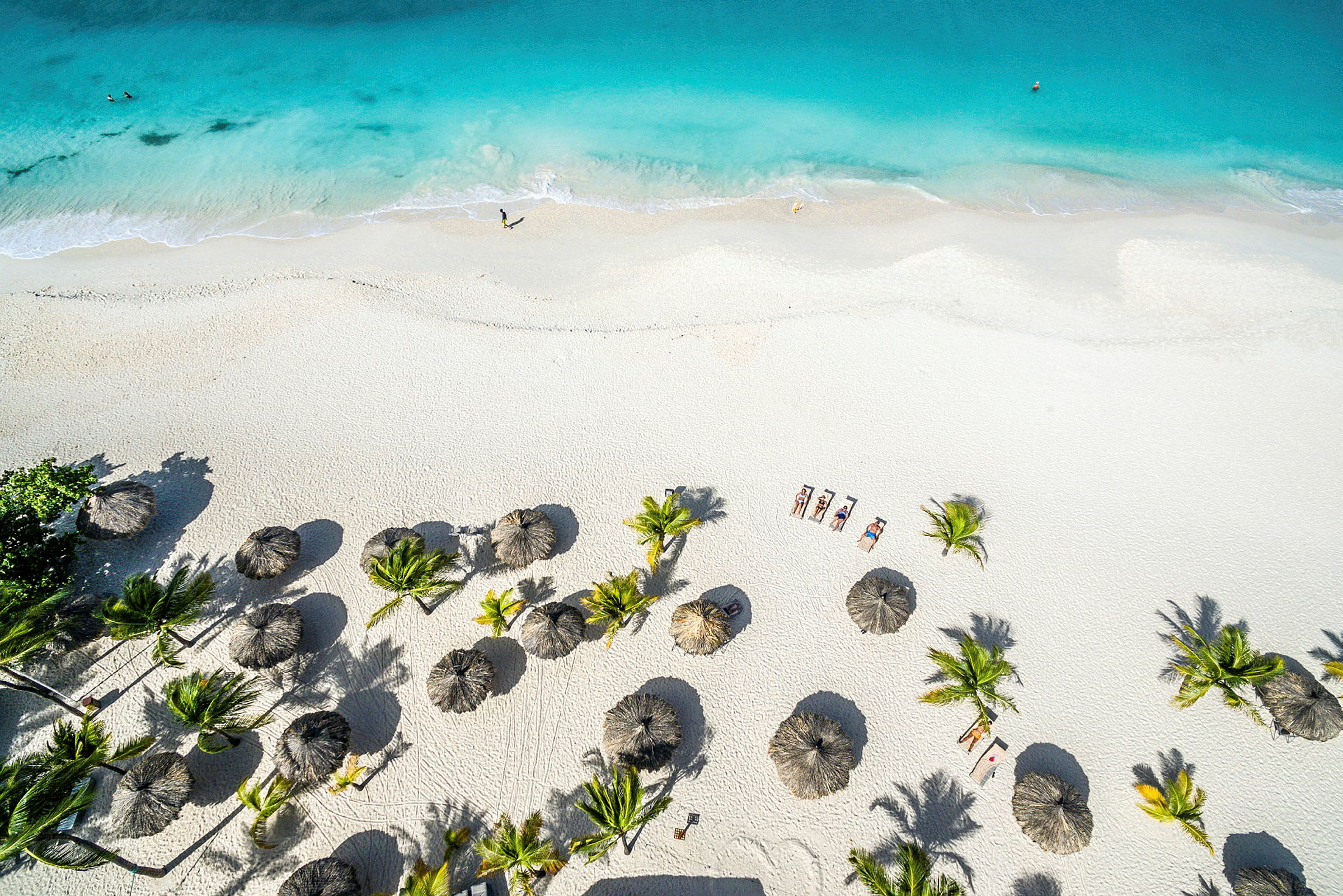 Luftfoto af en hvid sandstrand med turkisblåt vand. Stranden er dekoreret med parasoller og omgivet af palmer. (AI-genereret tekst)