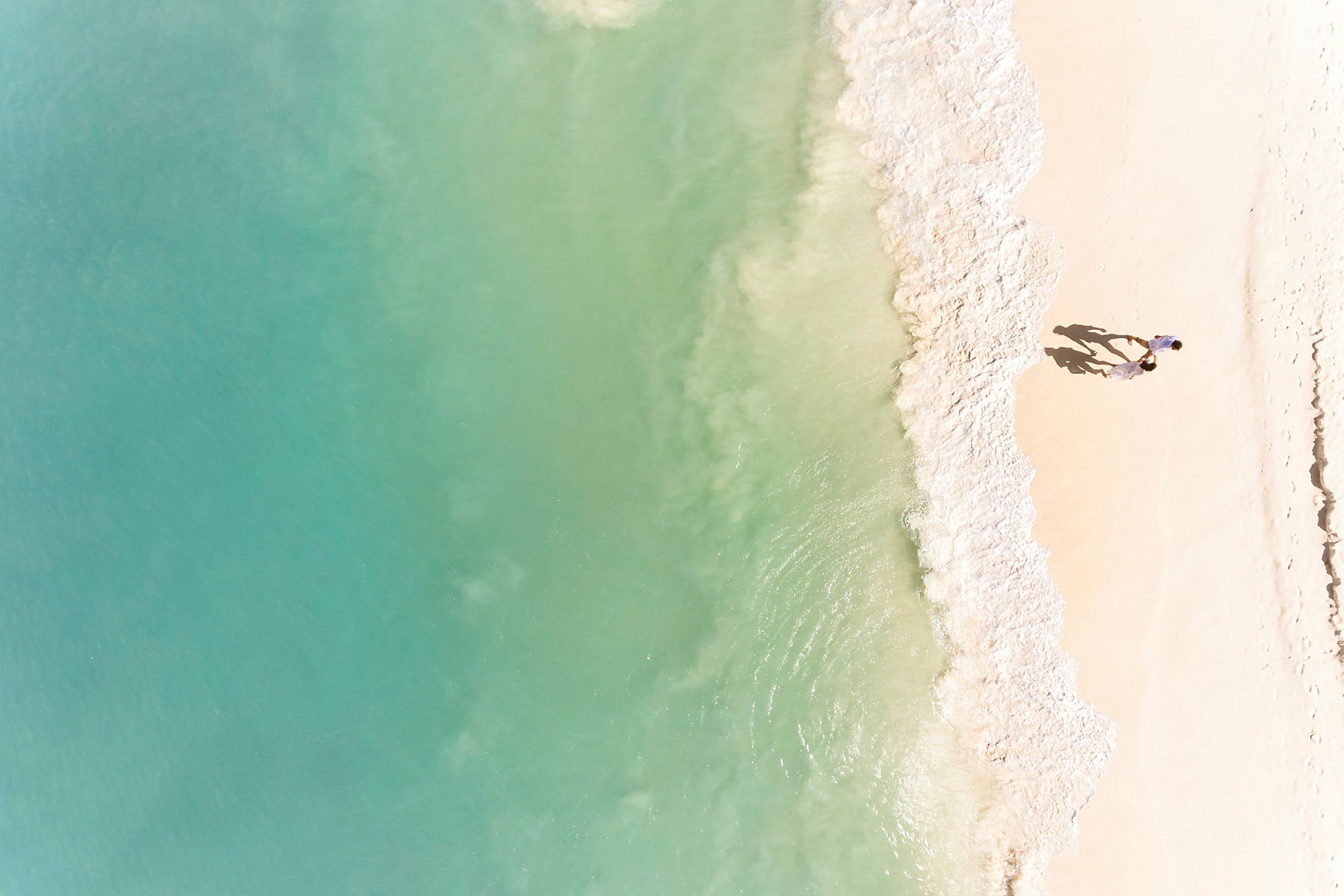 Et luftfoto af en strand med lyst sand, hvor bølger fra et turkis hav møder stranden. Et par personer går på stranden. (AI-genereret tekst)