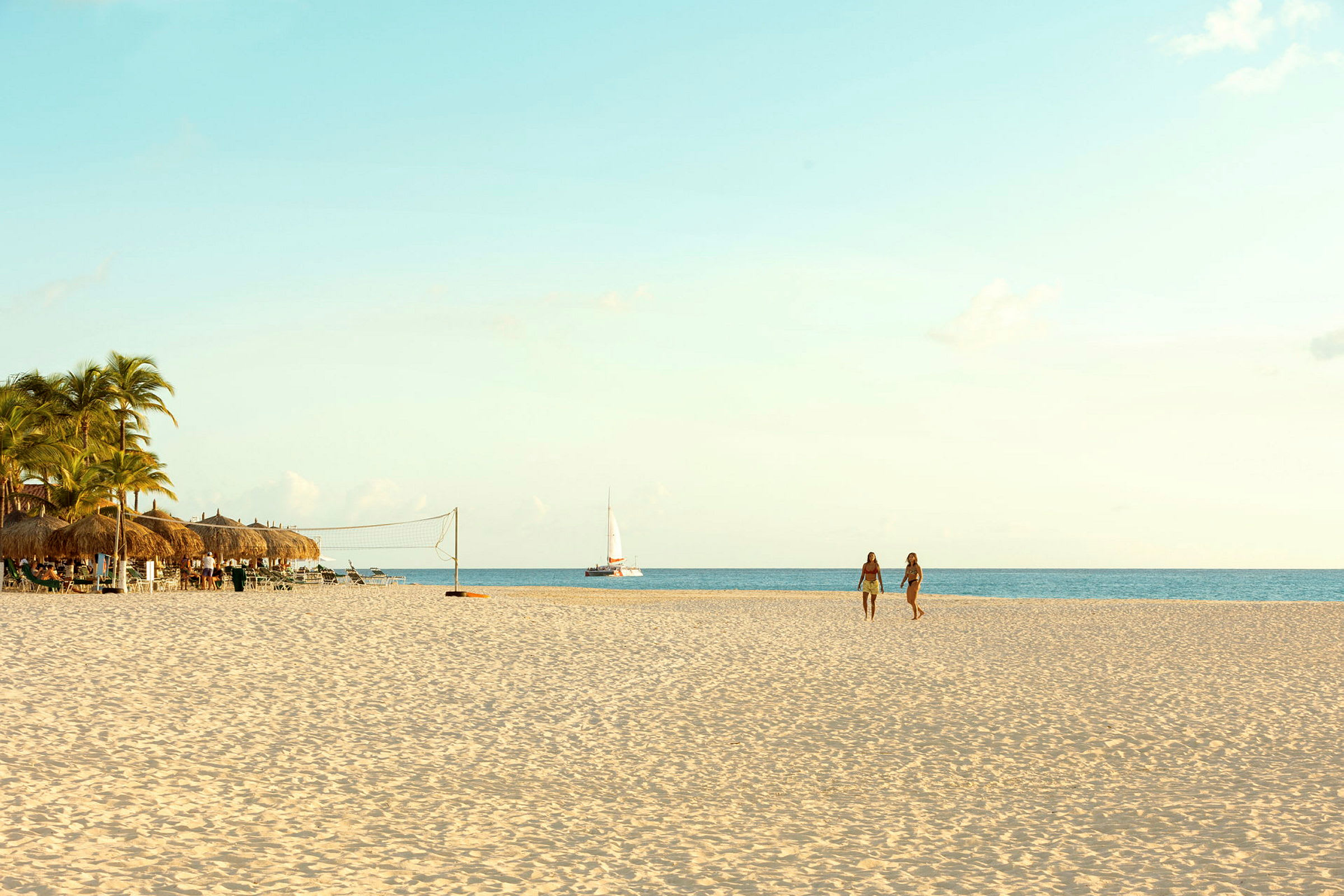 En sandstrand med palmer og parasoller til venstre, to personer der går på stranden og en sejlbåd på havet. (AI-genereret tekst)