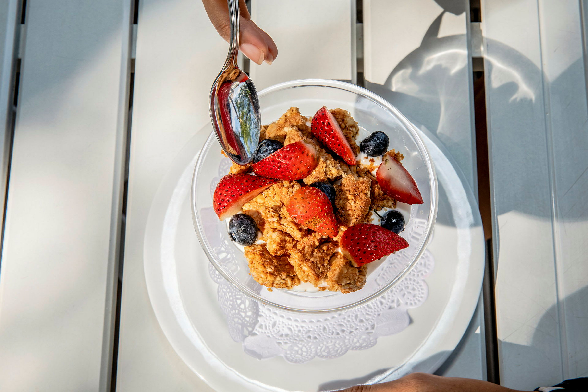 En skål med yoghurt, friske jordbær, blåbær og sprød granola. En hånd holder en ske over maden. (AI-genereret tekst)