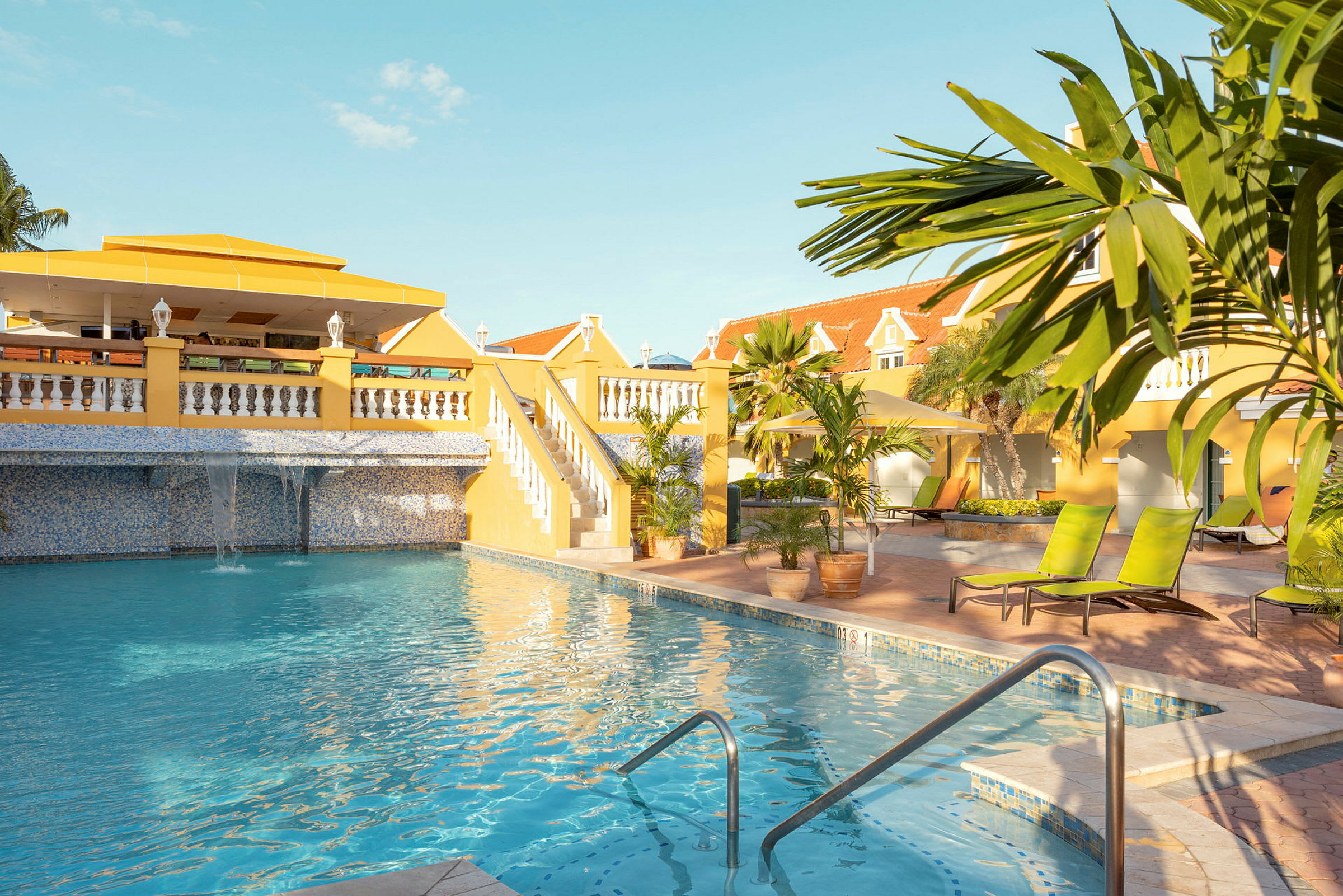 En udendørs swimmingpool med klart blåt vand omgivet af liggestole og palmer. I baggrunden ses en bygning med gul facade og røde tage. (AI-genereret tekst)