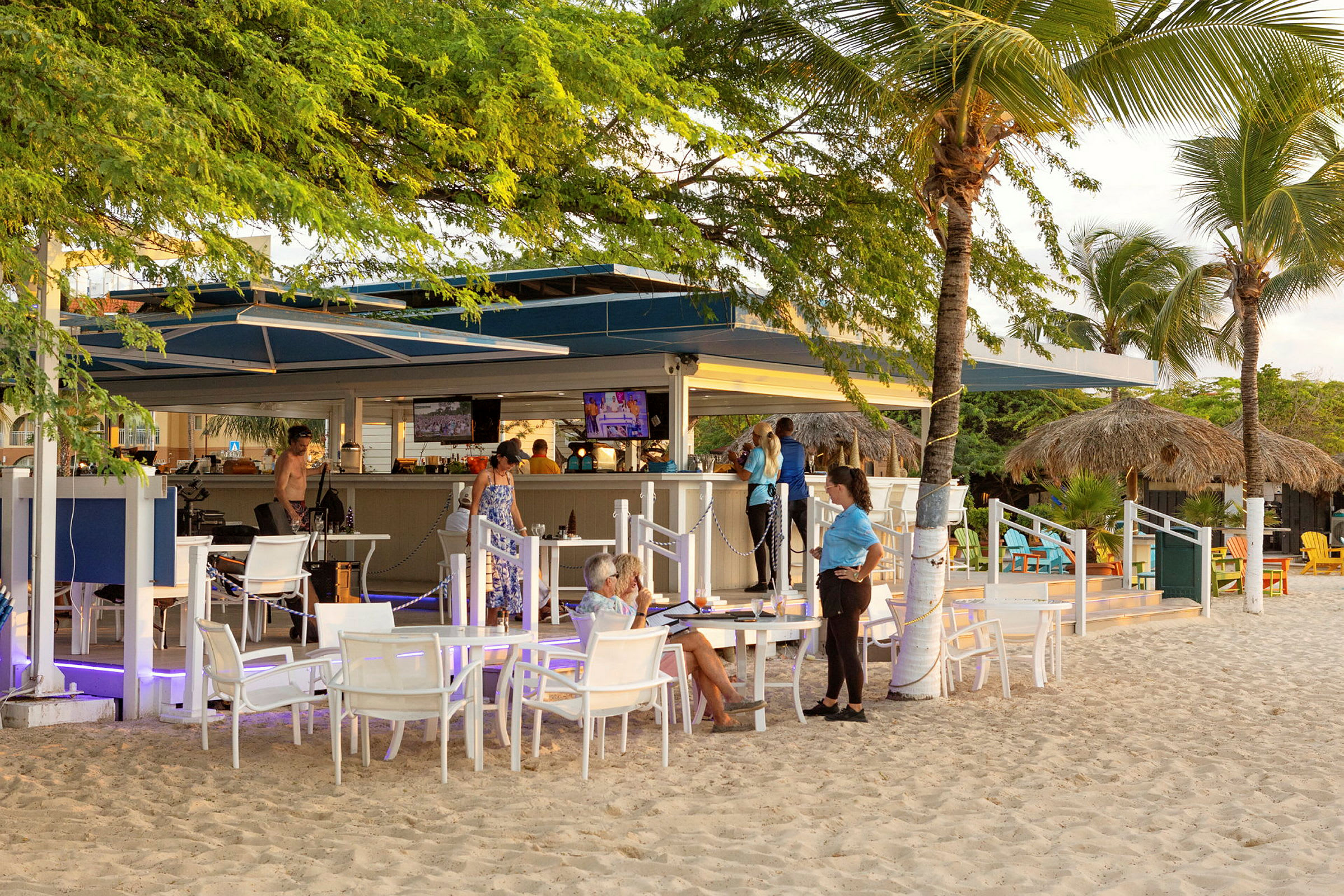 En strandbar med udendørs siddepladser på sanden, omgivet af vegetation og træer. (AI-genereret tekst)