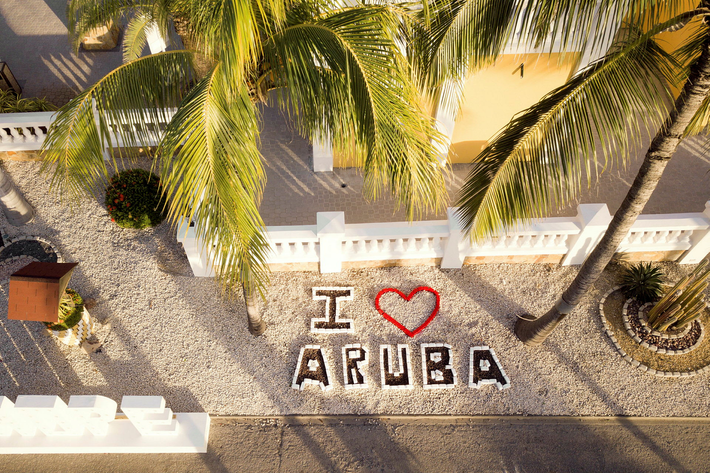 Teksten "I ❤️ Aruba" ses blandt dekorative stenbelægninger og palmer. (AI-genereret tekst)