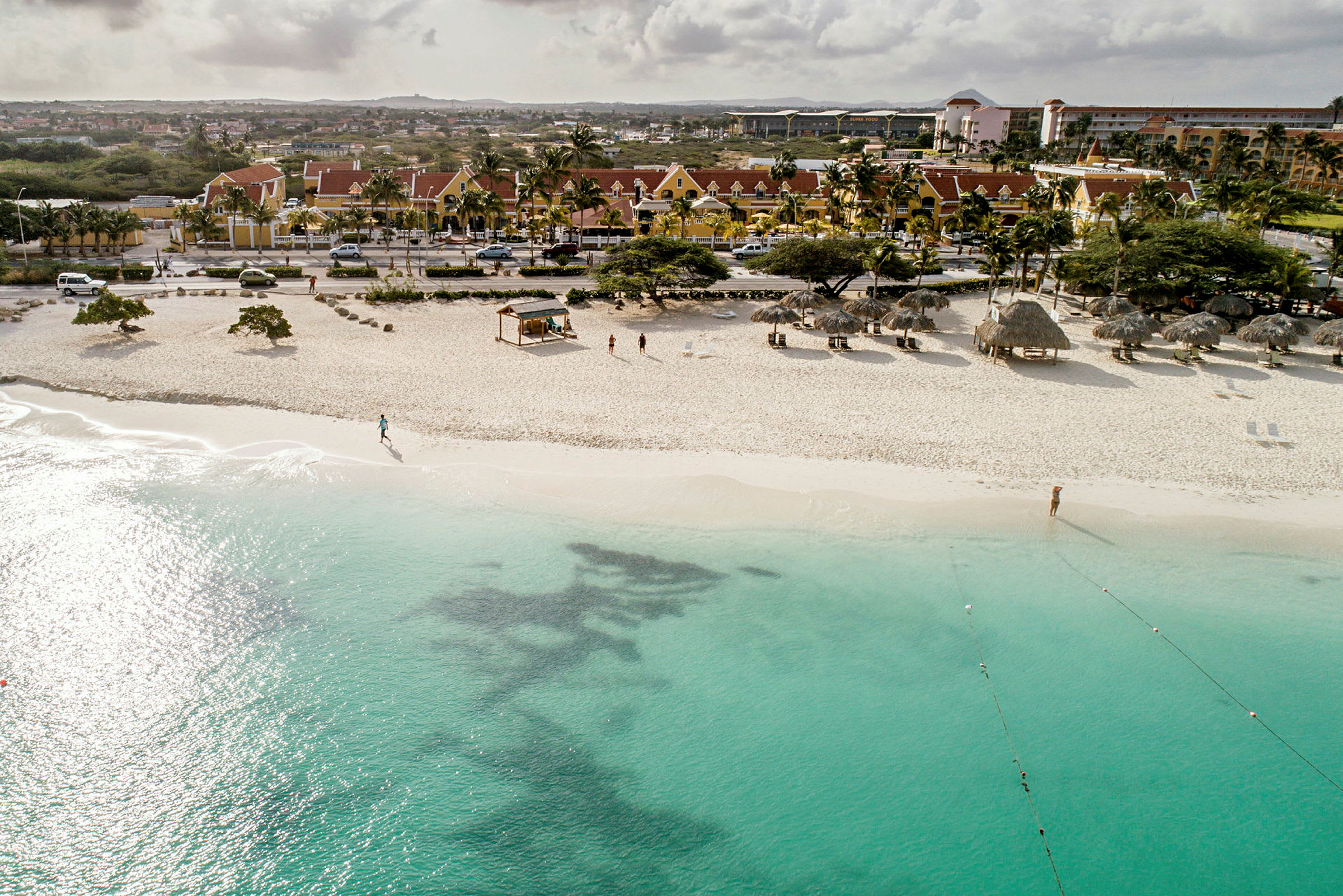 Sandstrand med turkis vand, parasoller og nogle personer på stranden, samt bygninger i baggrunden. (AI-genereret tekst)