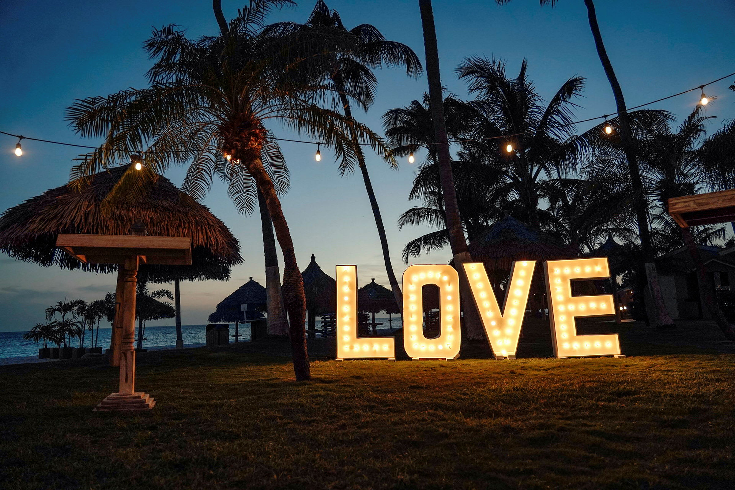 Et tropisk strandområde i skumringen med palmer, stråtag og store lysende bogstaver, der udgør ordet 'LOVE'. (AI-genereret tekst)