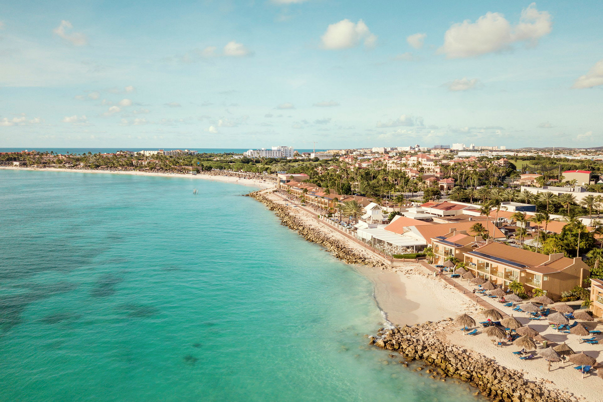 Strand med turkis vand, omkringliggende bygninger og solparasoller. (AI-genereret tekst)