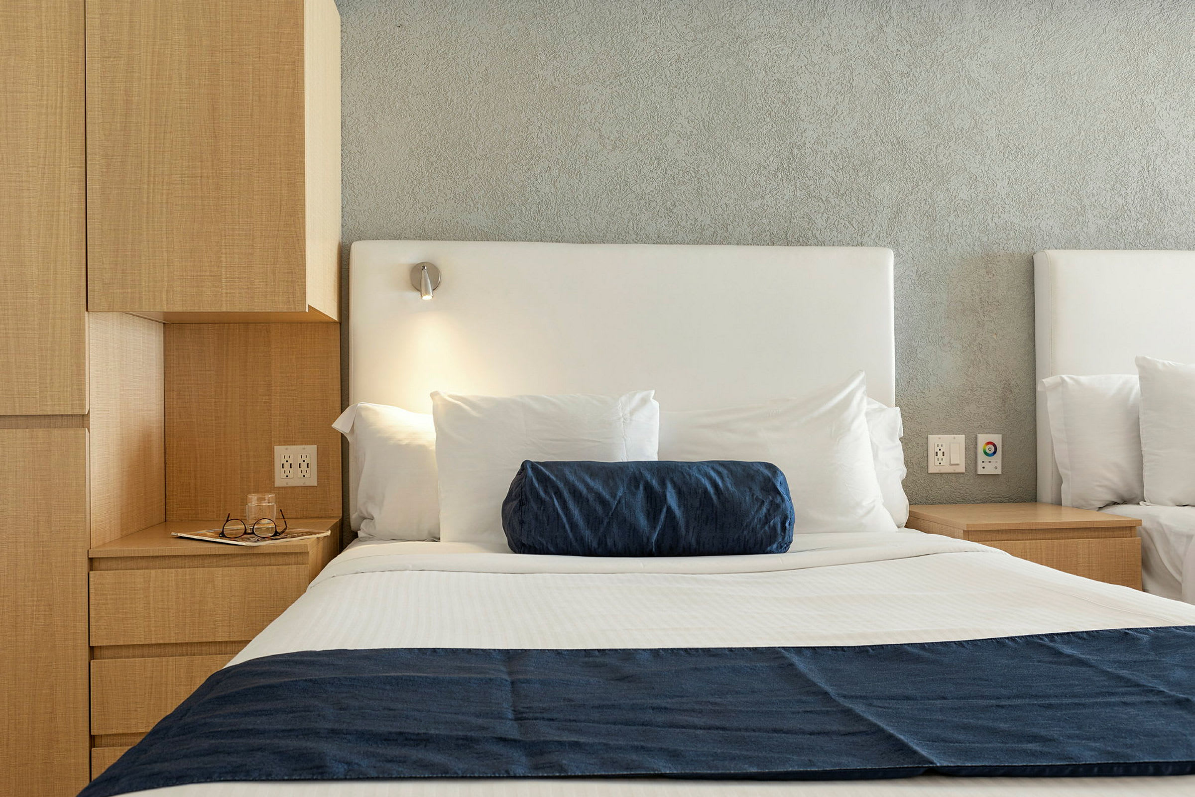 Billede af en hotelseng med hvidt sengetøj, en blå pyntepude og et blåt sengetæppe samt en sengelampe over. (AI-genereret tekst)