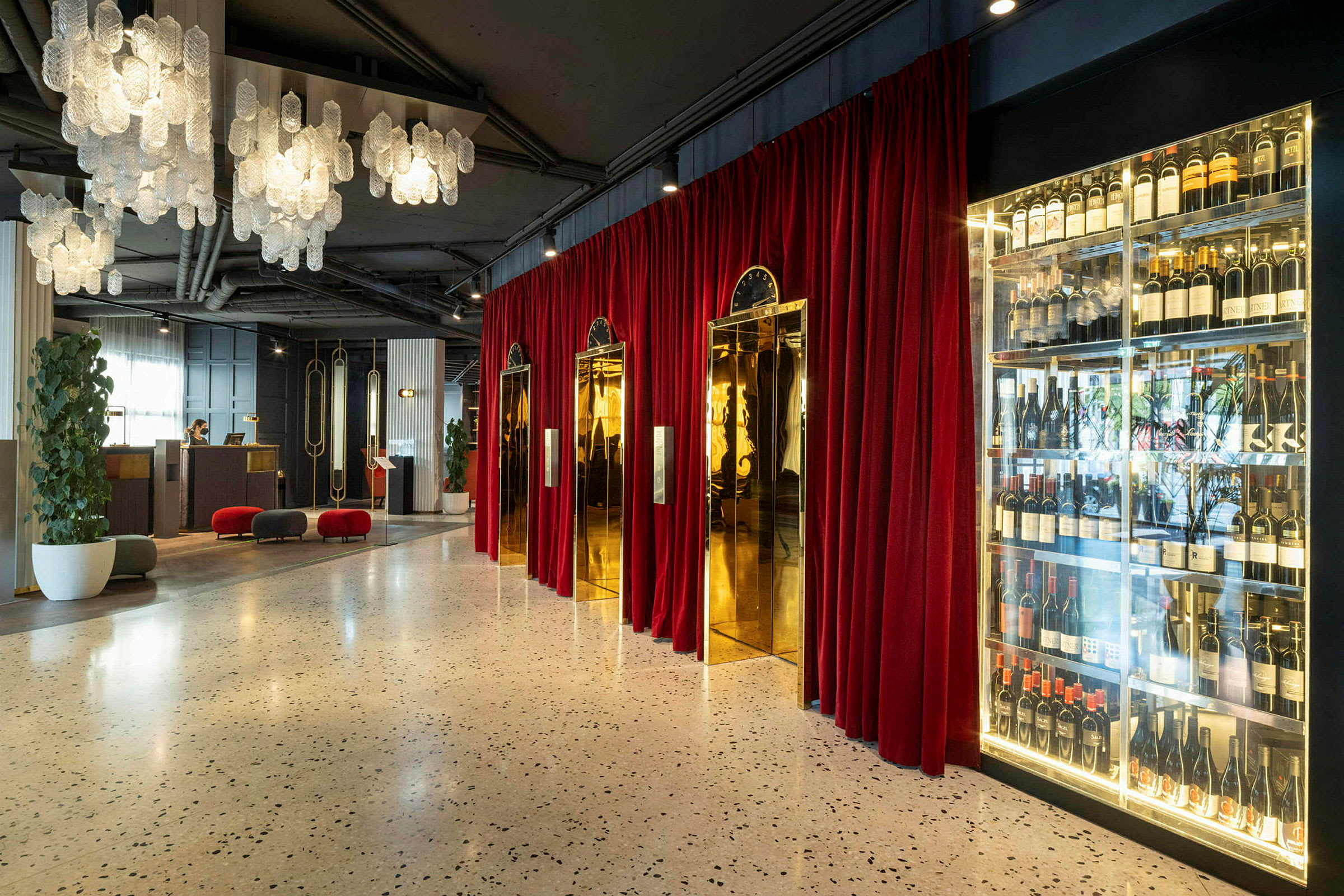 Hotellobby med hængende lysekroner, røde gardiner, gyldne elevatordøre og vinopbevaring. (AI-genereret tekst)