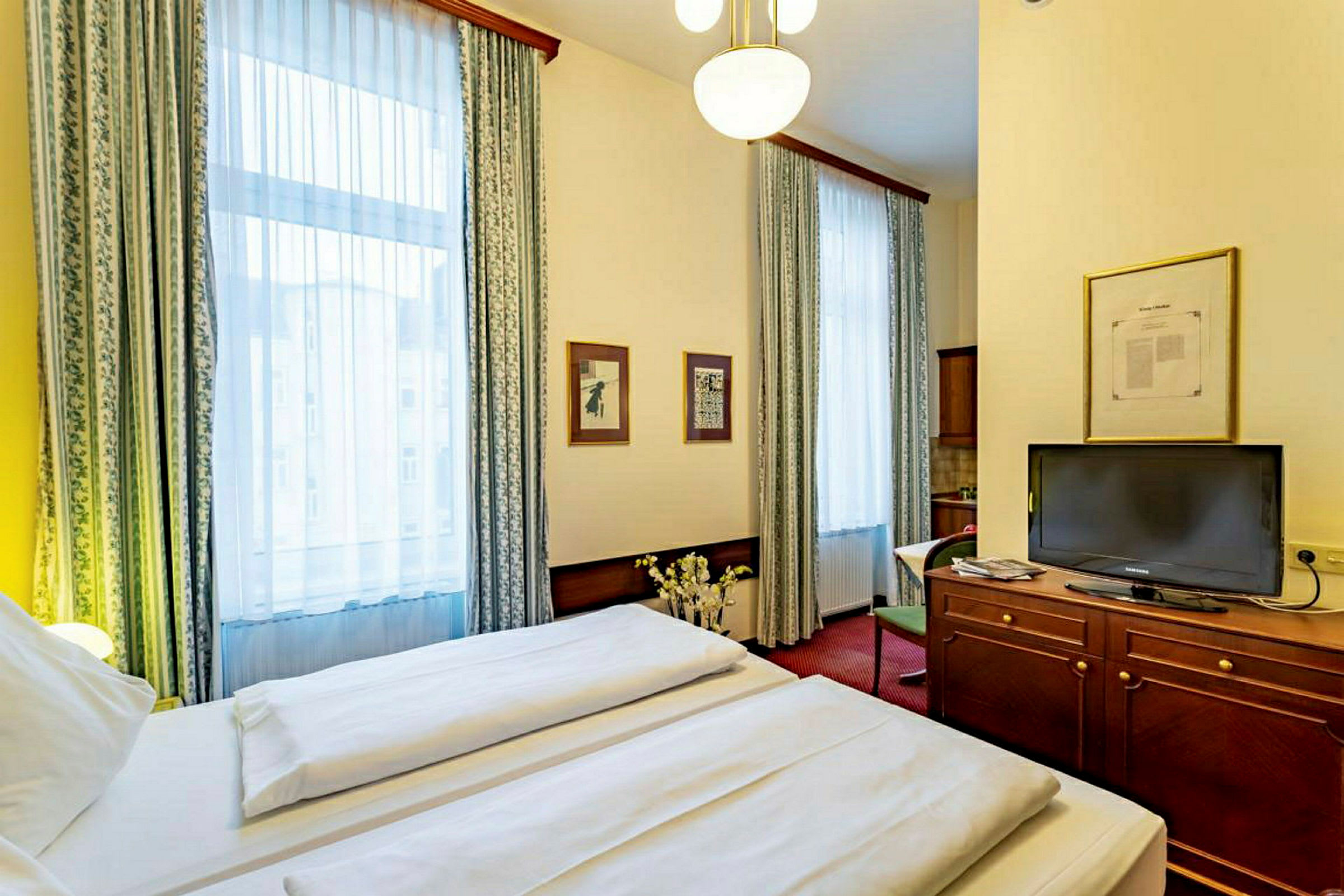 Interiør af et hotelværelse med en dobbeltseng, et TV på en trækommode og to store vinduer med gardiner. (AI-genereret tekst)