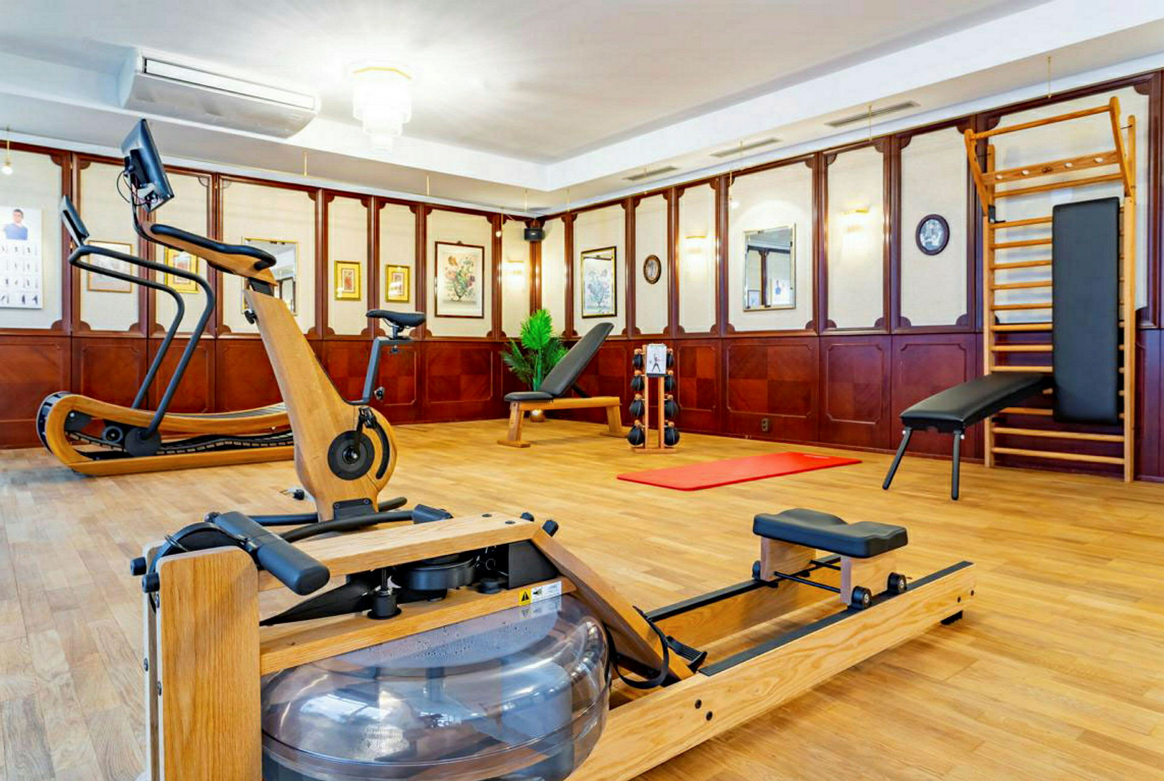 Billede af et træningsrum med forskellige fitnessmaskiner såsom romaskine, motionscykel og træningsbænk. Rummet har træpaneler og kunst på væggene. (AI-genereret tekst)
