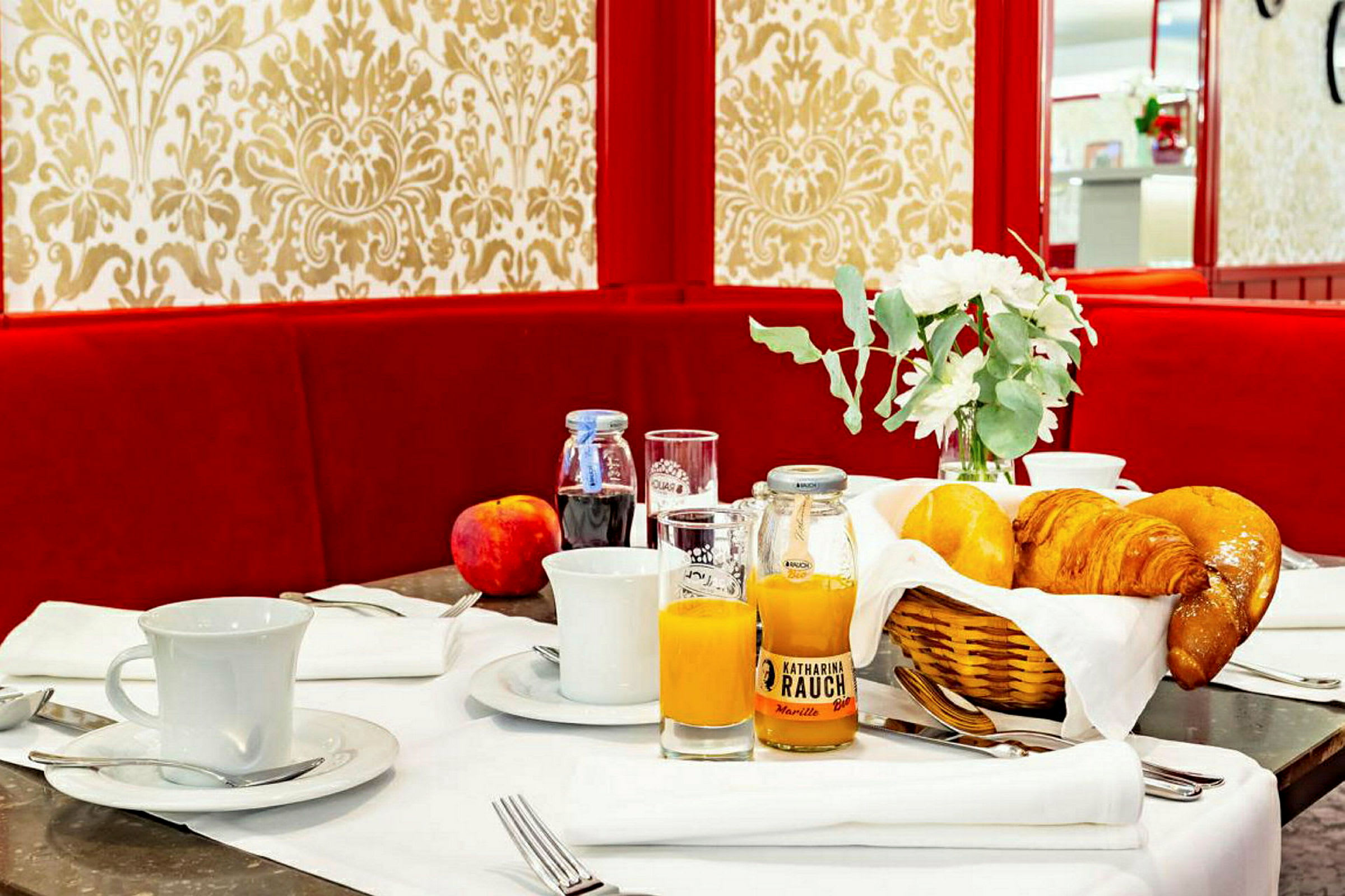 Morgenbord med bagværk, juice, kaffekopper og dekorationer mod en rød sofa og mønstret baggrund. (AI-genereret tekst)