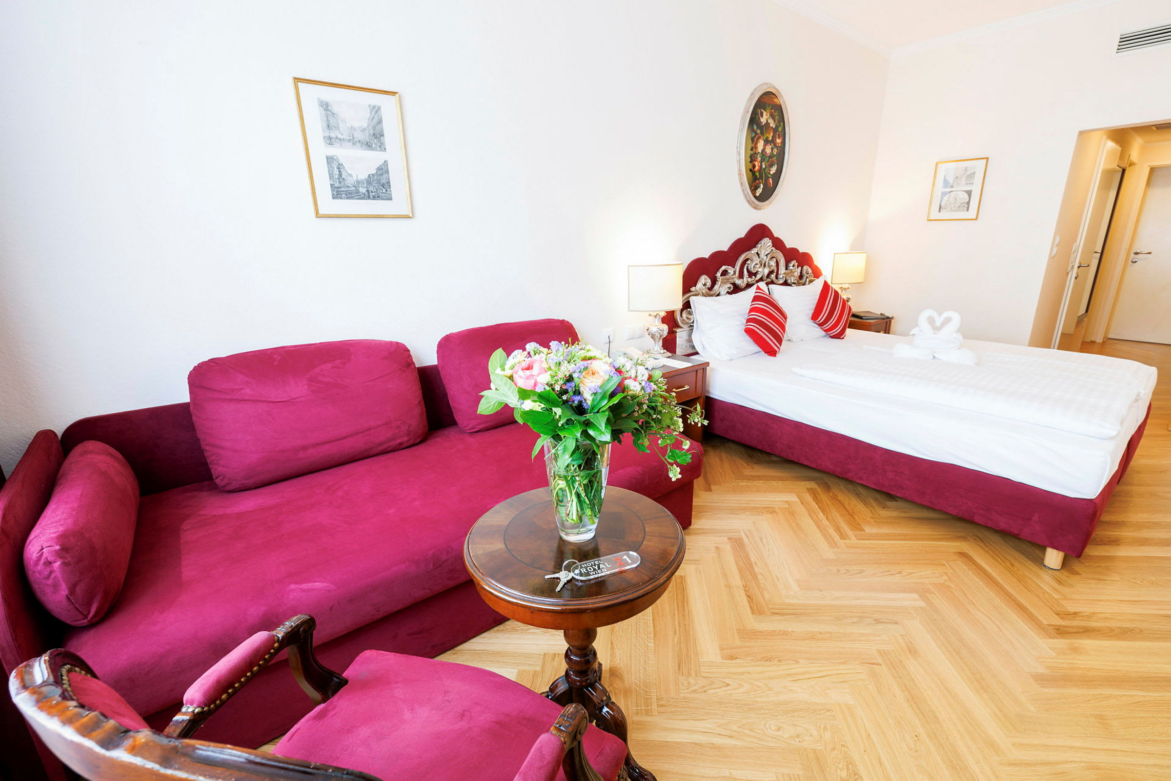 Et hotelværelse med en rød sofa, et rundt bord med en blomsterbuket og en seng med røde detaljer. (AI-genereret tekst)