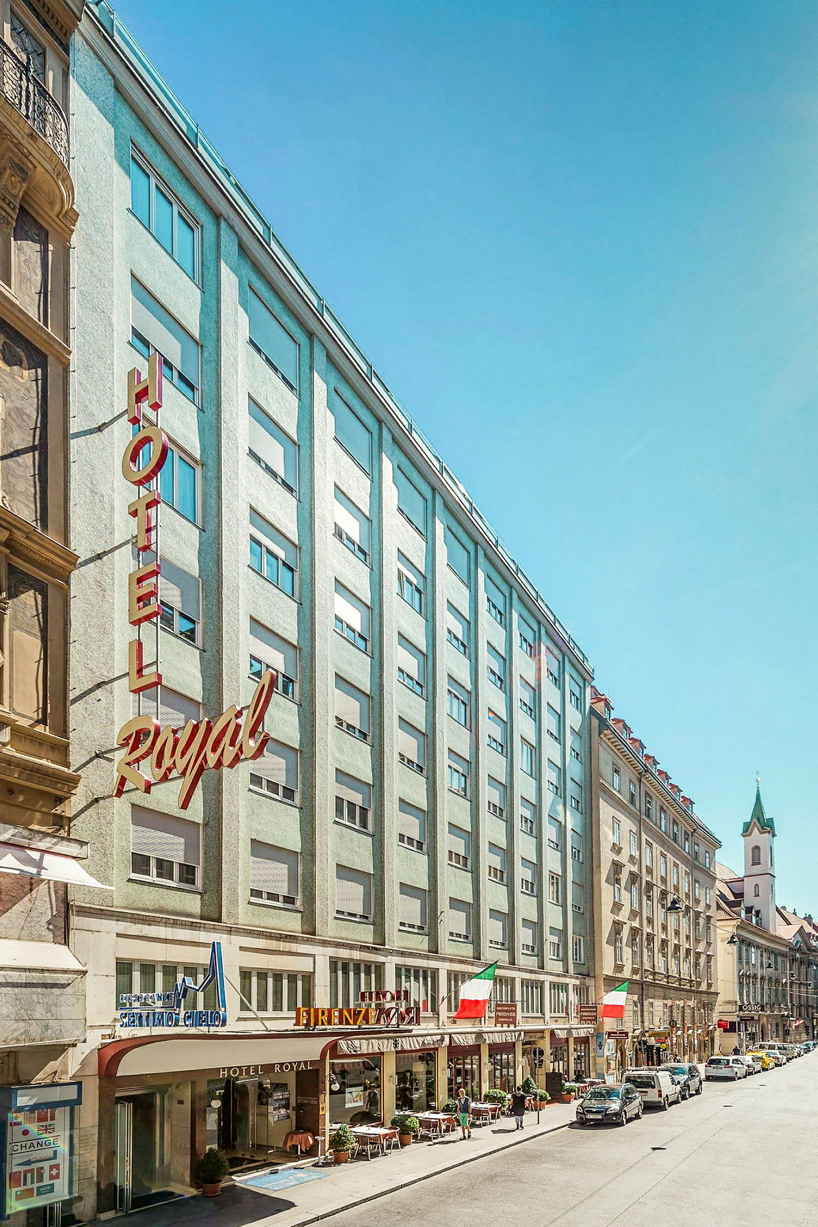Billede af en bygade med hotellets facade og omkringliggende bygninger. (AI-genereret tekst)