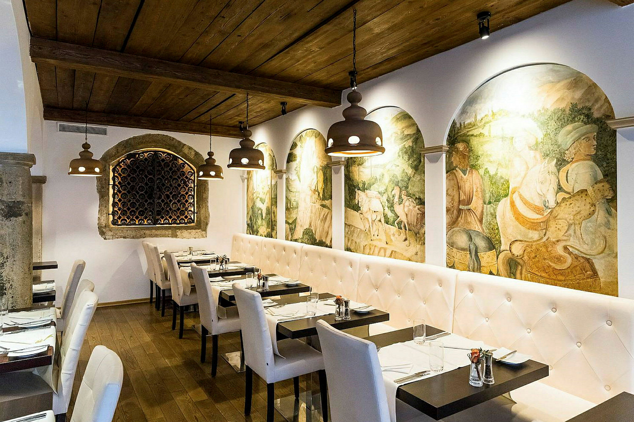 En restaurantindretning med hvide polstrede sofaer og stole, mørke træborde og en væg dekoreret med historiske freskomalerier. Loftet er af træ, og belysningen består af hængende armaturer. (AI-genereret tekst)
