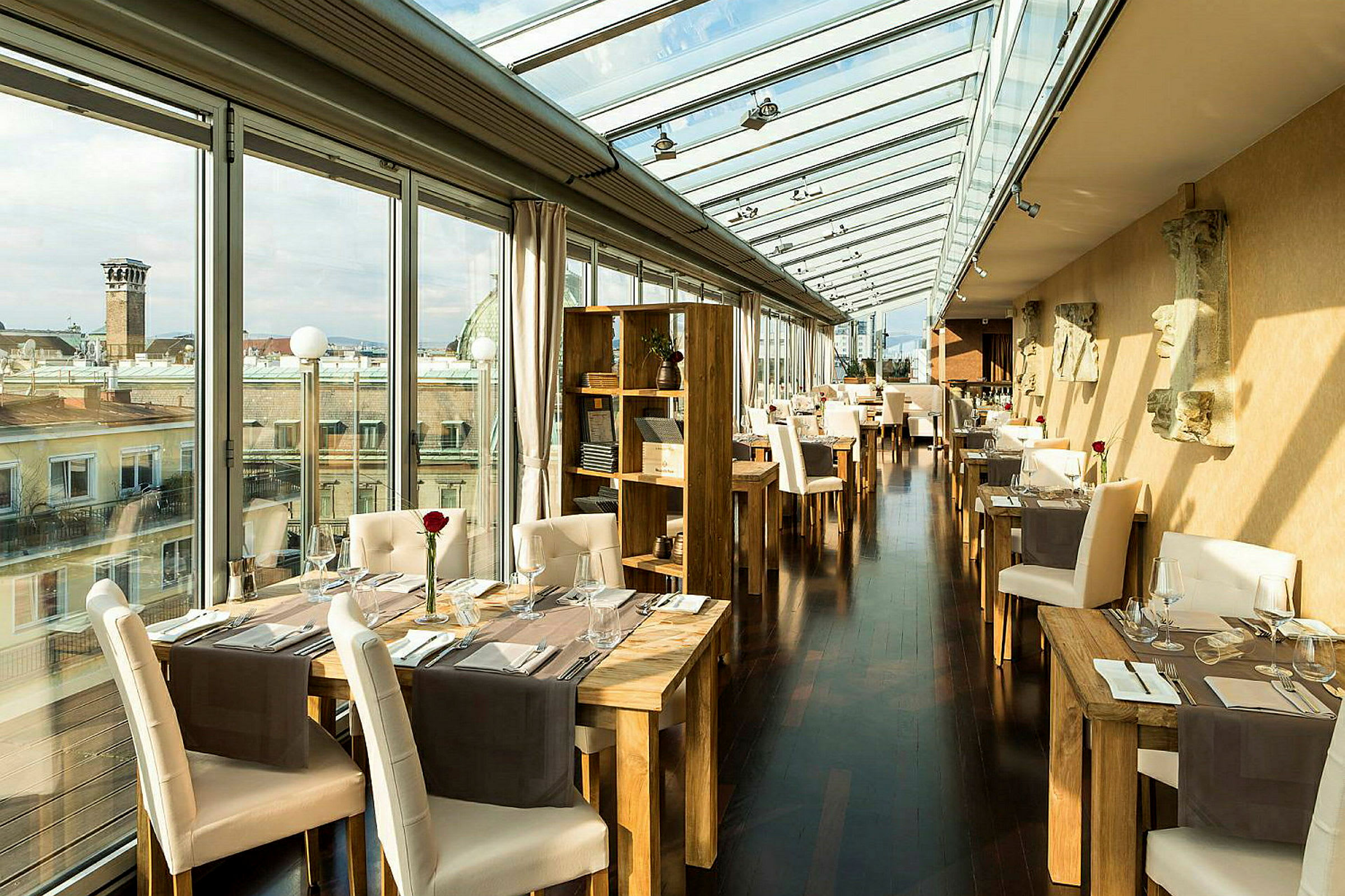 Et elegant restaurantområde med store glasvægge, der giver udsigt over byens hustage. Bordene og stolene er elegant arrangeret med røde roser i glasvaser som borddekoration. (AI-genereret tekst)