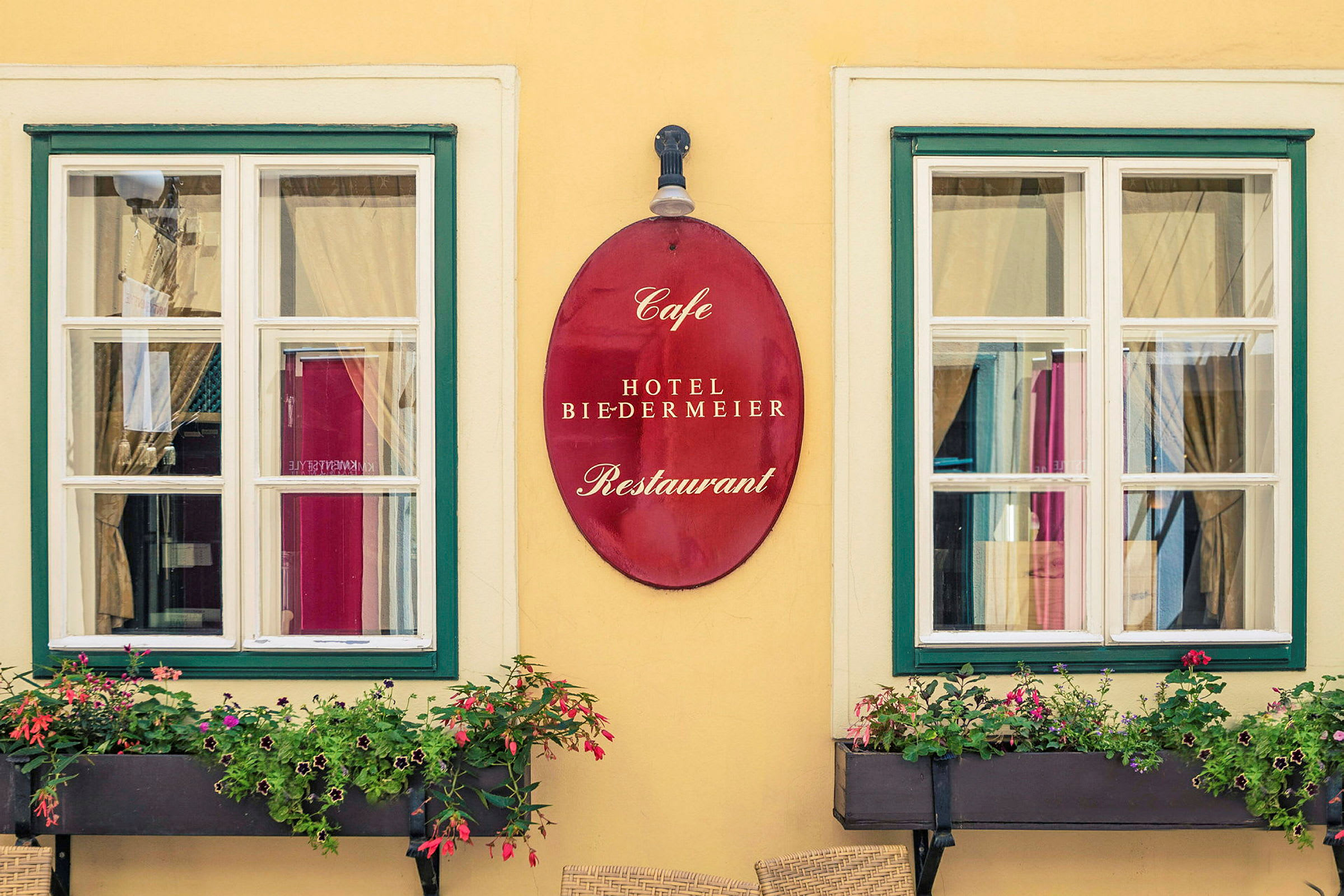 To vinduer med hvide karme og grønne rammer, mellem dem hænger et rødt skilt med teksten "Cafe Hotel Biedermeier Restaurant". Foran vinduerne er der blomsterkasser med blomstrende planter. (AI-genereret tekst)