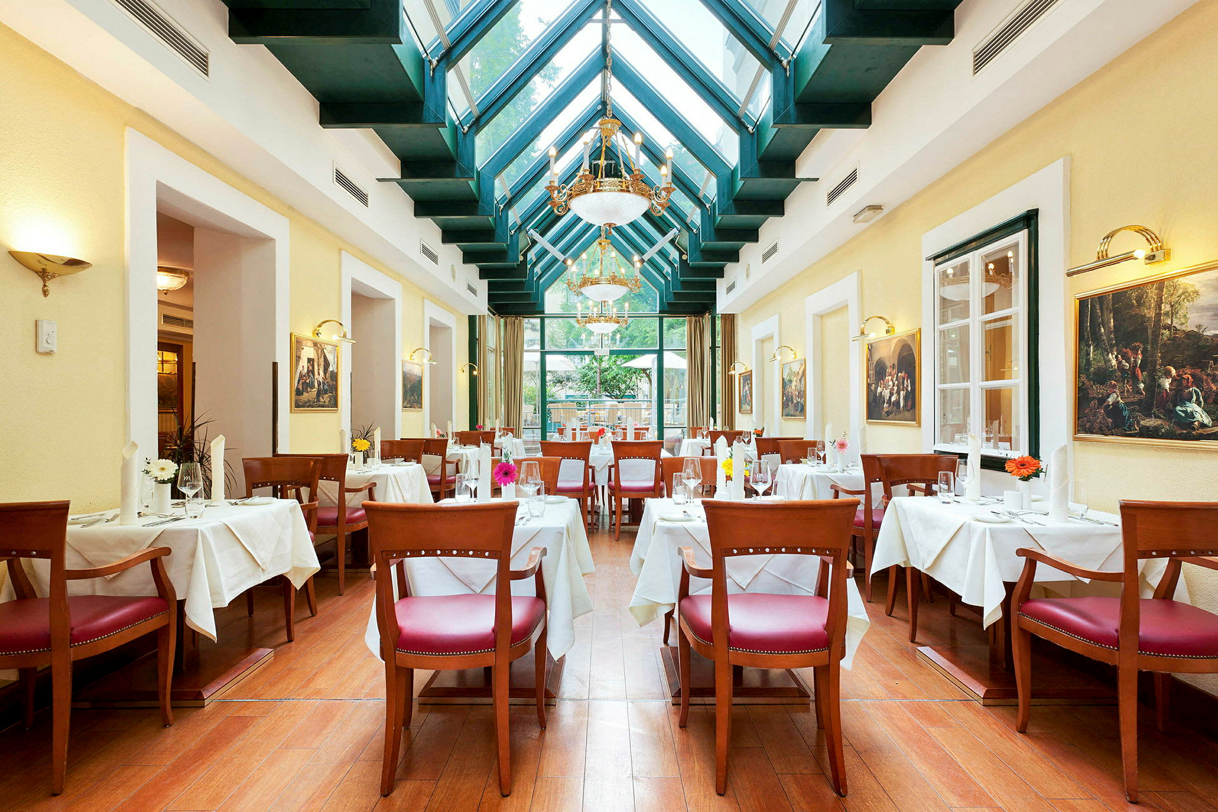 En elegant restaurant med højt loft og glastag. Bordene er dækket med hvide duge, og stolene har røde sæder. Væggene er dekoreret med malerier. (AI-genereret tekst)