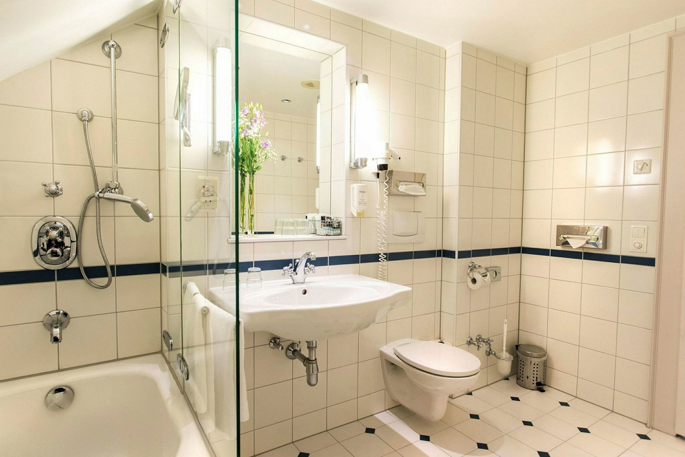 Et badeværelse med hvide fliser, badekar, bruser, vask, spejl og toilet. (AI-genereret tekst)
