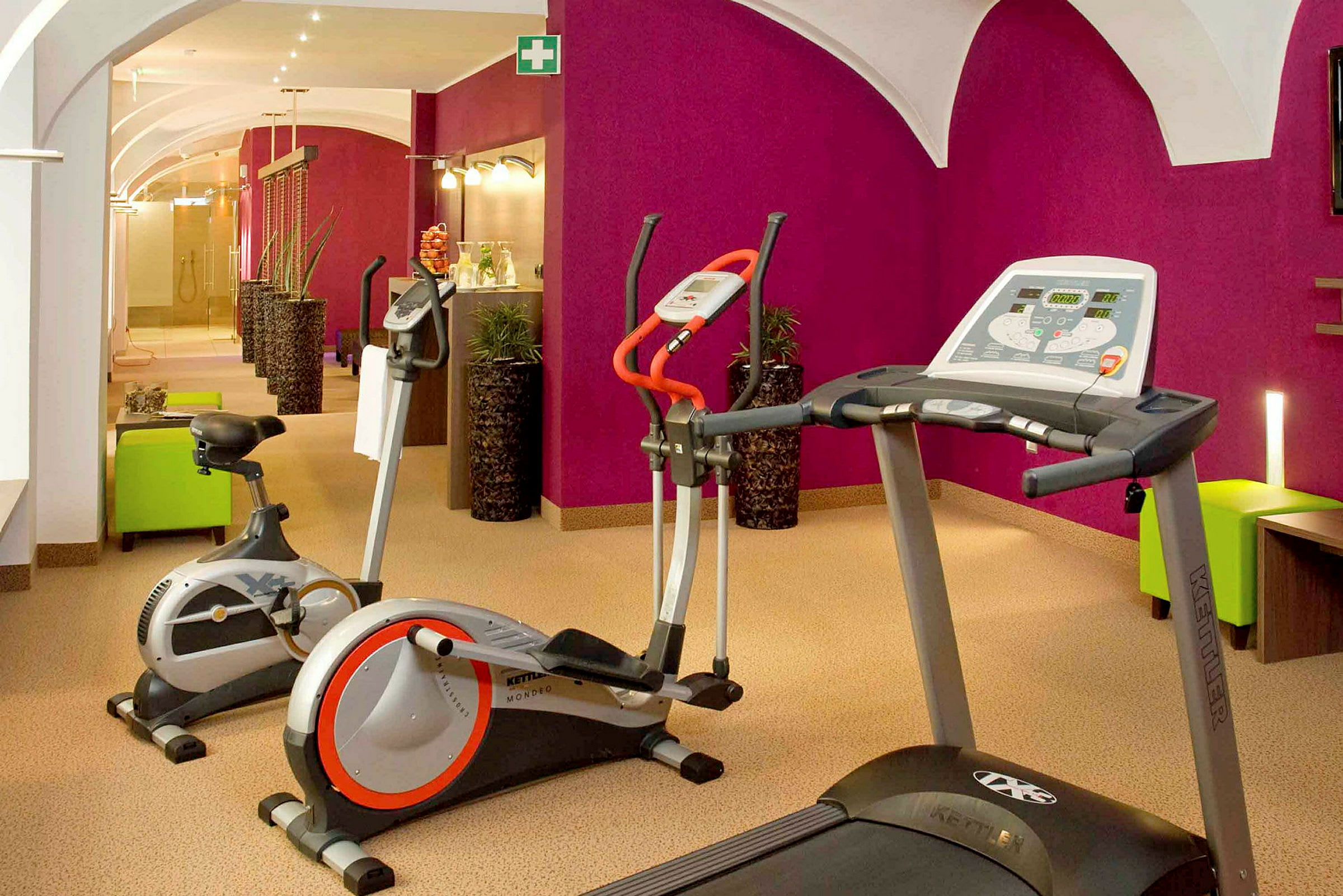 Et fitnessrum med træningsmaskiner, herunder løbebånd og motionscykler, placeret i et rum med lilla vægge og lys indretning. (AI-genereret tekst)