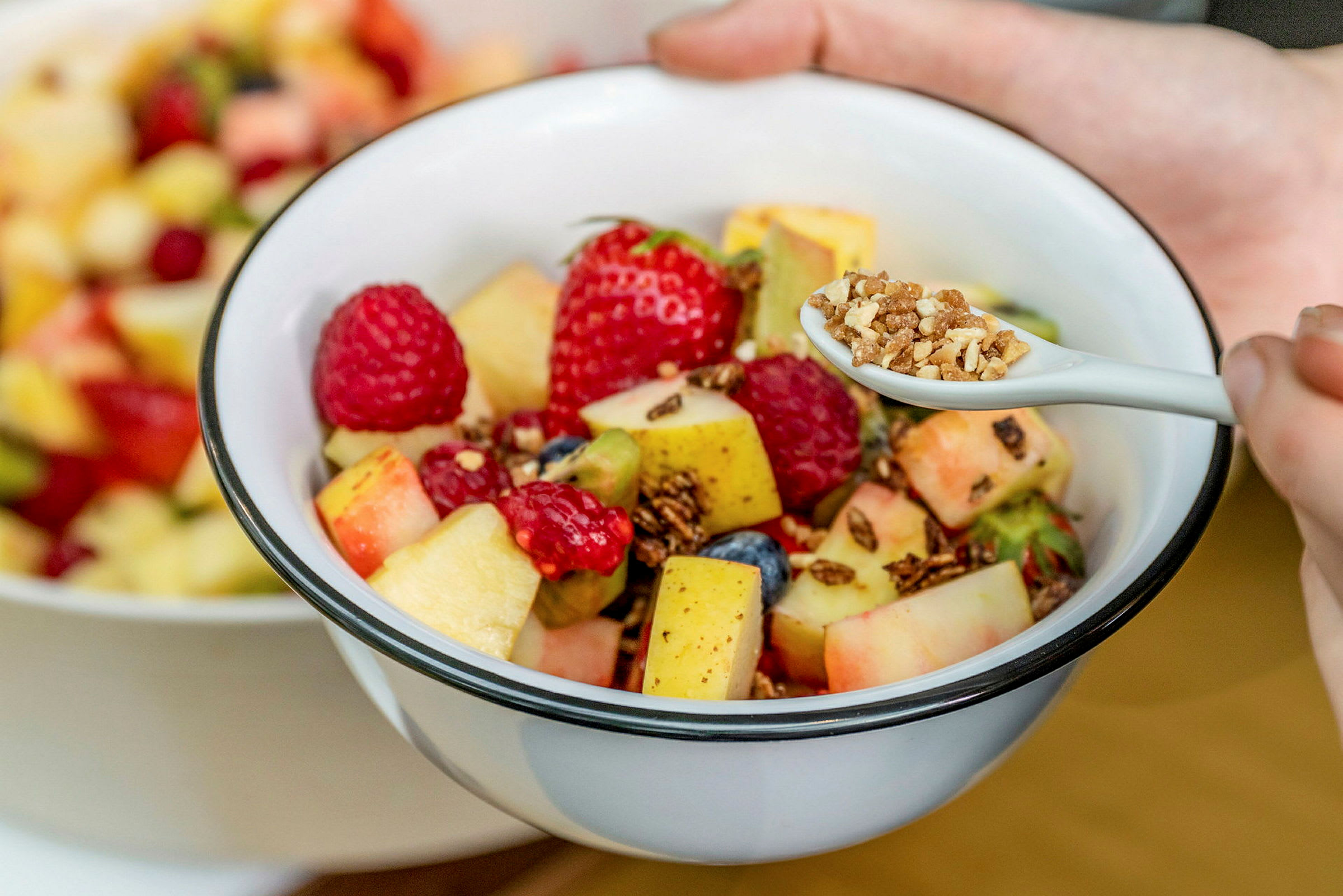 En skål med frugtsalat bestående af forskellige frugter og bær, samt en ske med granola. (AI-genereret tekst)