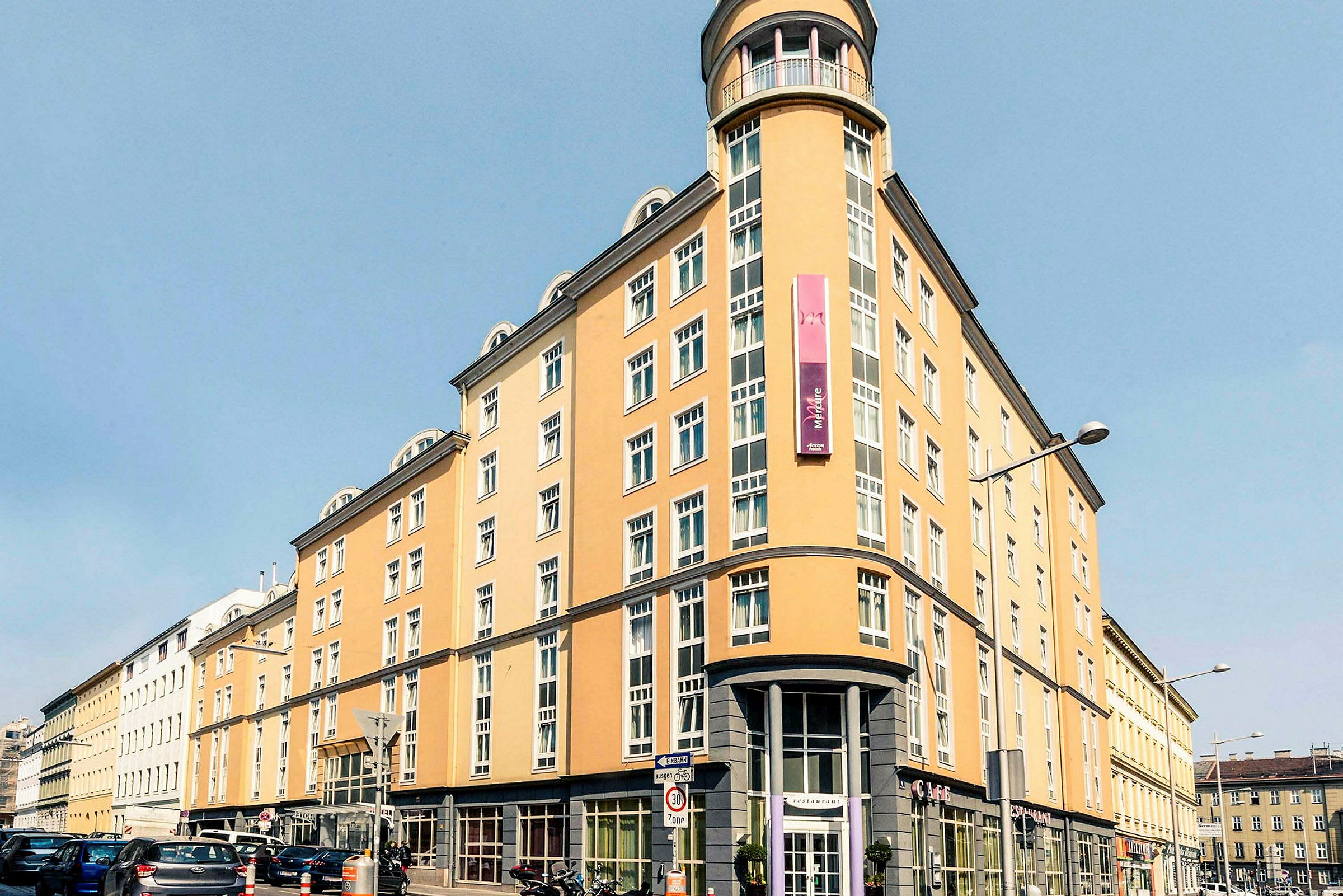 En lysegul bygning med mange vinduer, et skilt og afrundet hjørnefacade. (AI-genereret tekst)