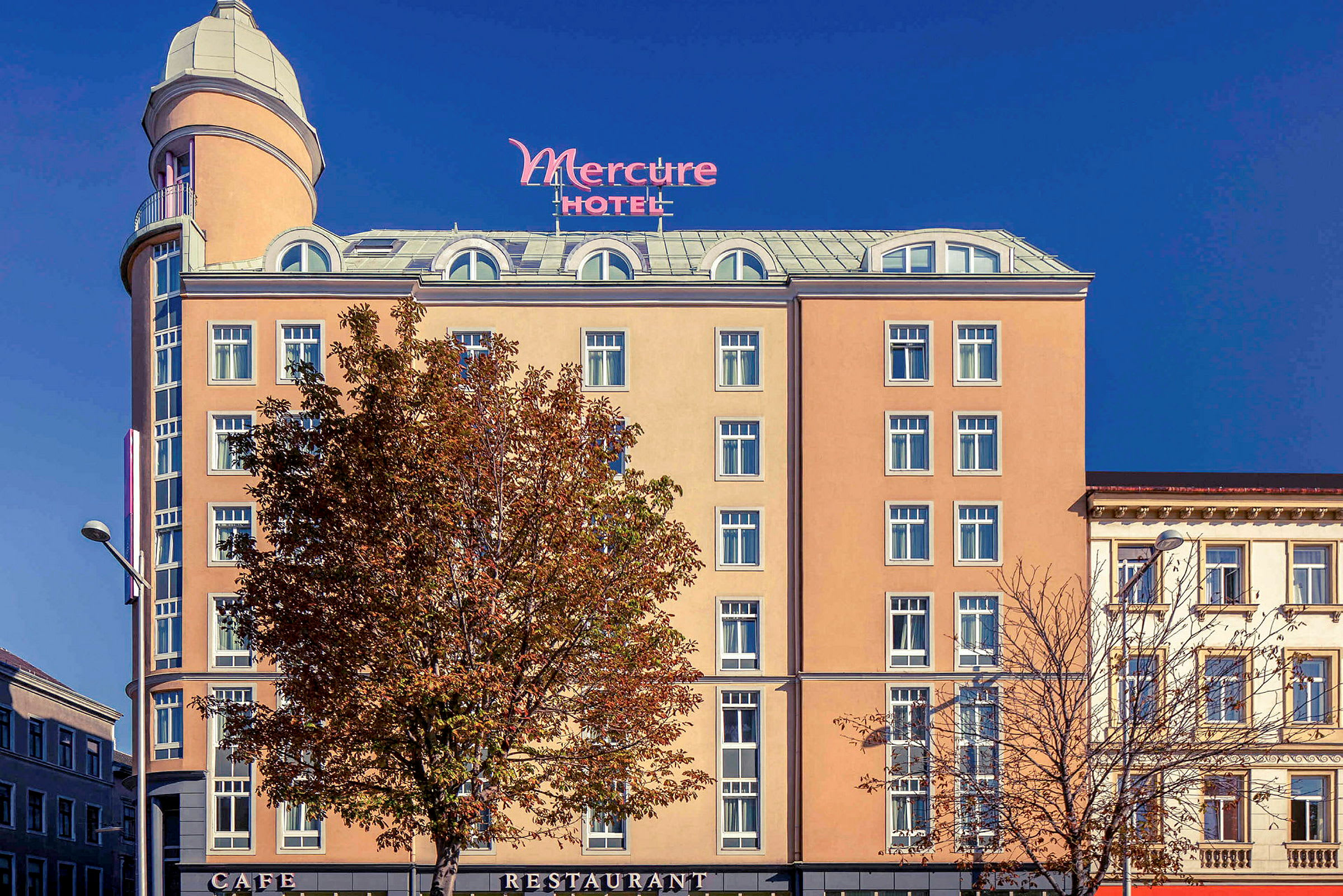 Eksteriøret af et hotel med navnet Mercure, med en beige facade, mange vinduer og et højt tårn øverst. (AI-genereret tekst)