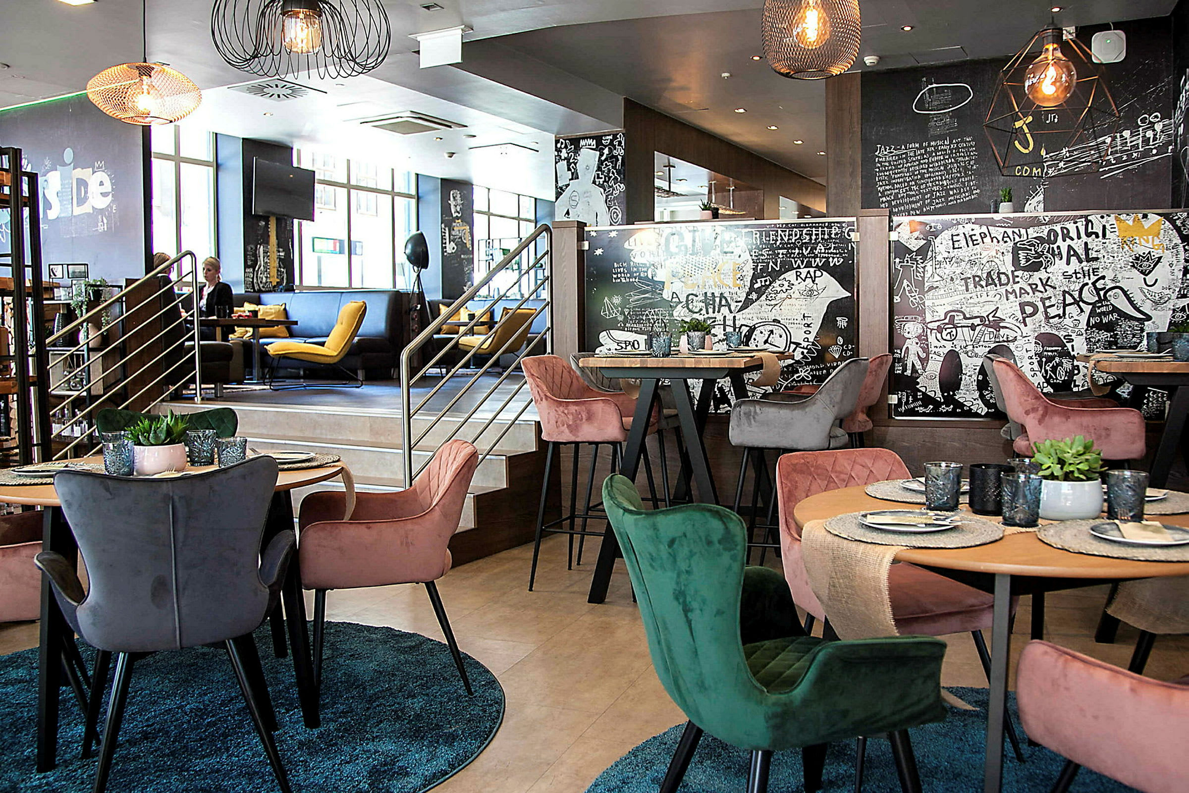 En moderne indretning af en restaurant med farverige stole omkring borde med dækkede tallerkener og glas. Væggene er dekoreret med kunstneriske mønstre og tekster. (AI-genereret tekst)