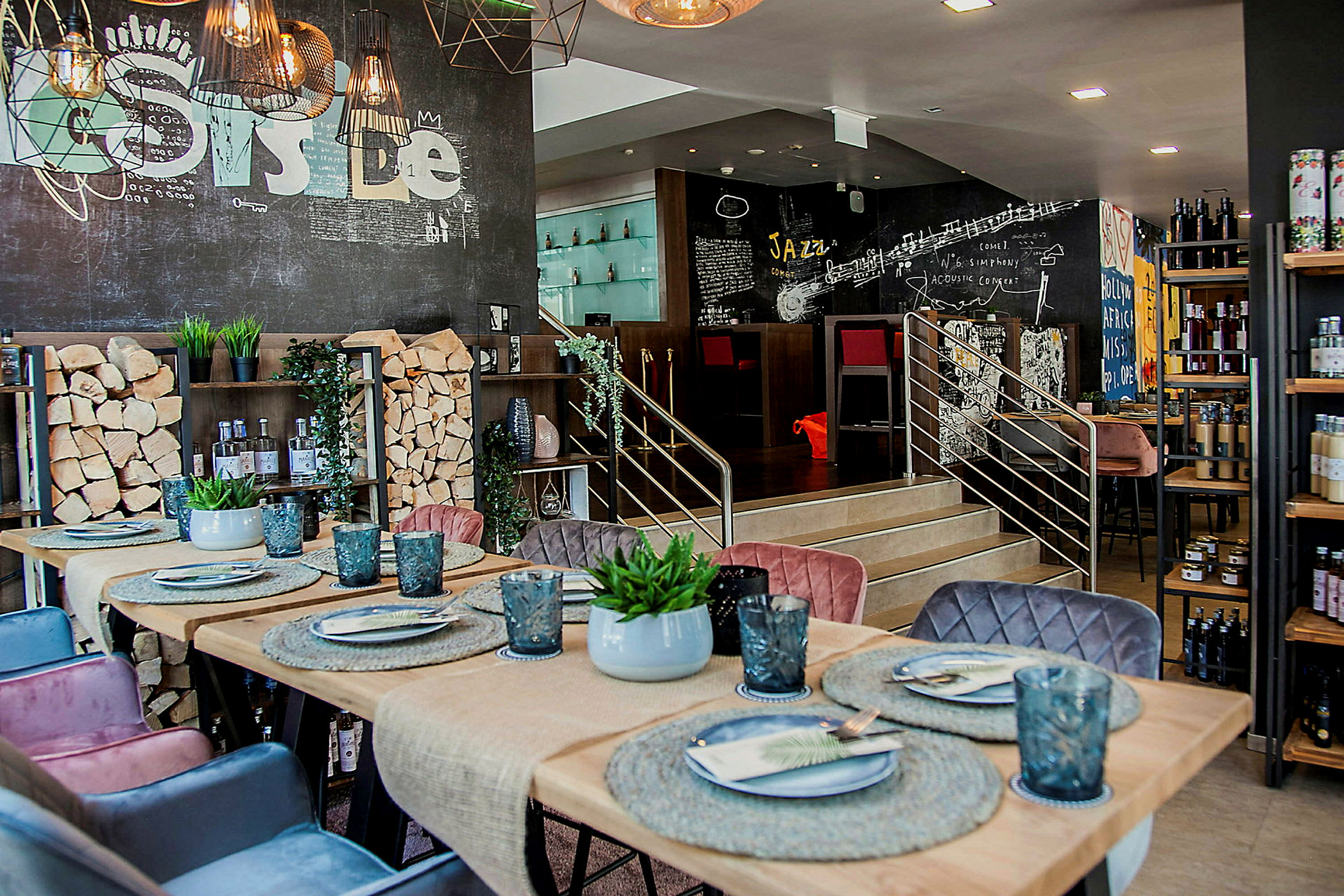 En restaurant eller café med moderne interiørdesign. Bordet er dekoreret med tallerkener, glas og en lille plante. Væggene har kreative malerier, og der er stakke af træ som en del af indretningen. (AI-genereret tekst)