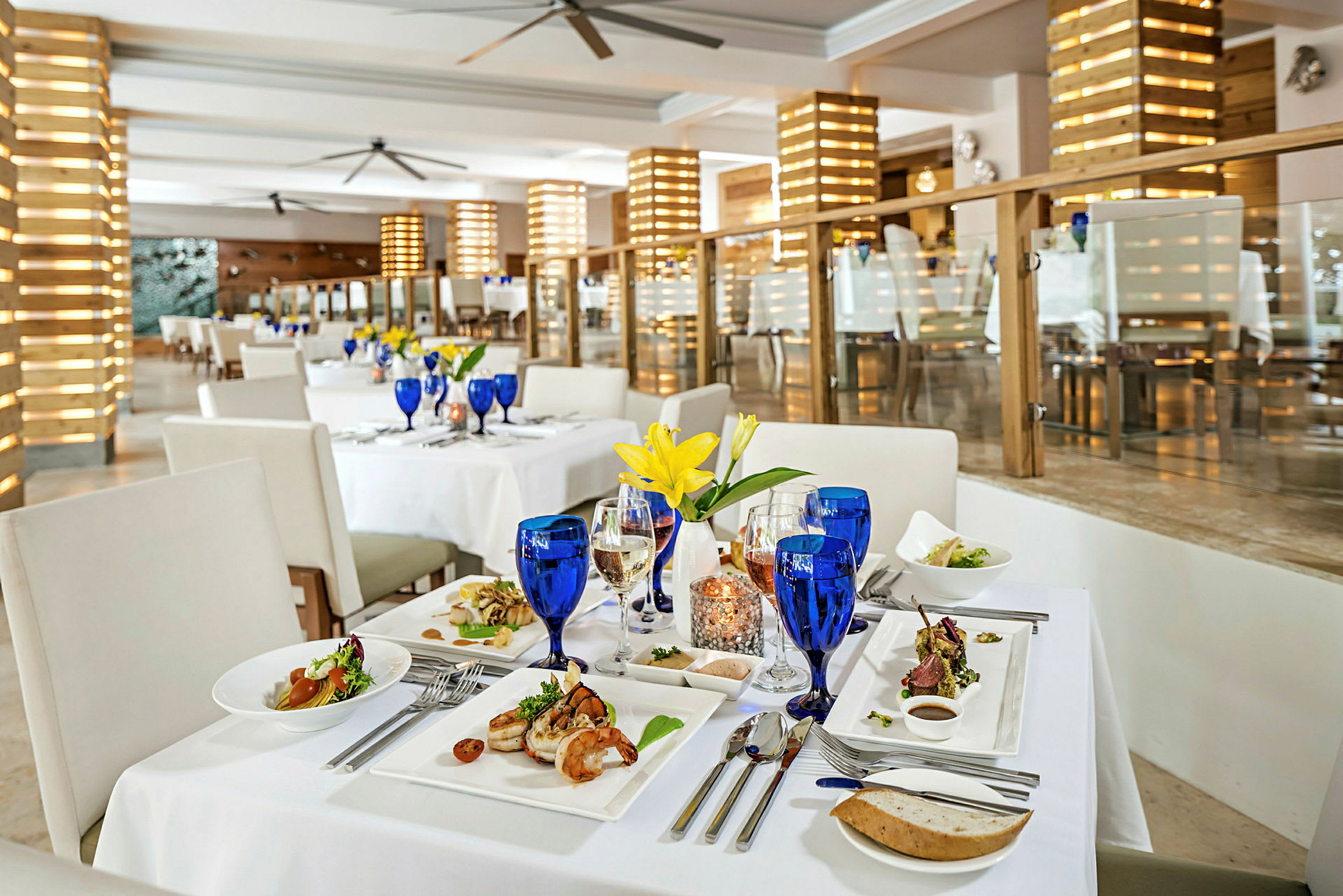 Restaurantbord med hvid dug, blå glas og tallerkener med mad. (AI-genereret tekst)