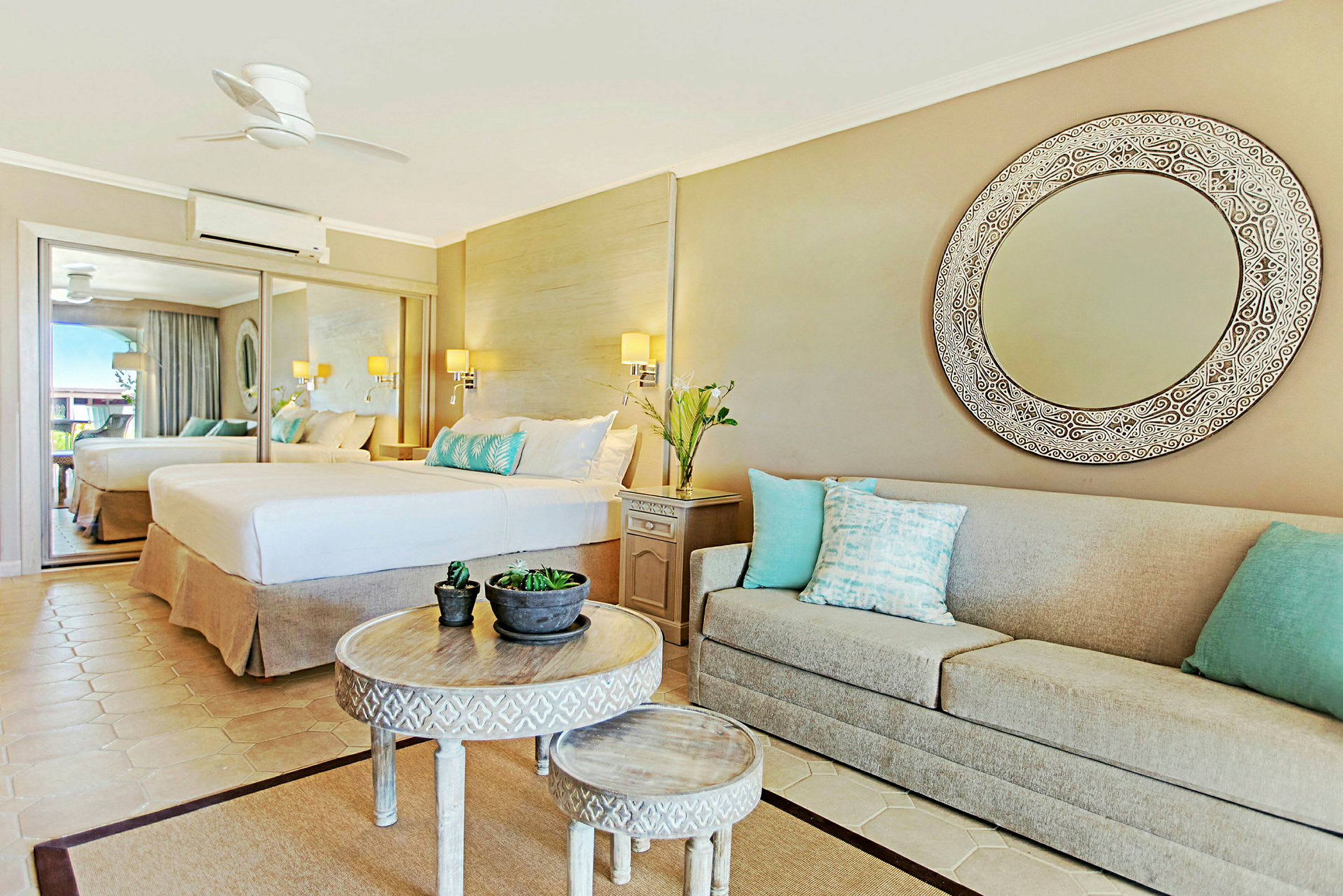 Et elegant hotelværelse med et stort spejl, en bred seng og en beige sofa med dekorative puder. Et rundt bord med mindre bord og plantepynt i midten. (AI-genereret tekst)