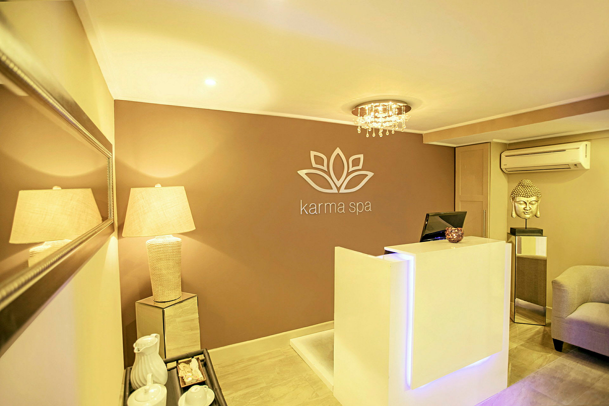 En reception i et spa-anlæg med et moderne og elegant design. En hvid receptionsdisk står foran en væg med logo, og en lampe oplyser rummet. (AI-genereret tekst)