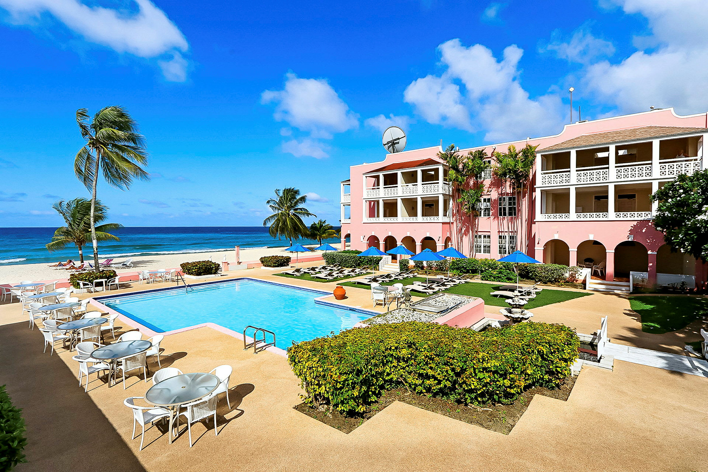 Billede af en solrig ferieby med en udendørs pool, omkringliggende terrasser og et lyserødt hotel med altaner, beliggende ved havet. (AI-genereret tekst)
