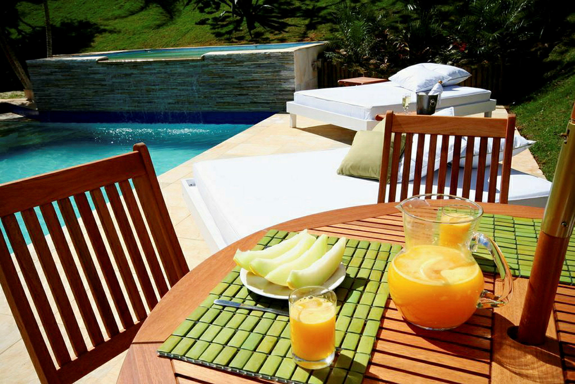 Billedet viser et bord ved en pool med en kande og et glas juice samt en tallerken med udskåret frugt. I baggrunden ses solsenge og grønne planter. (AI-genereret tekst)