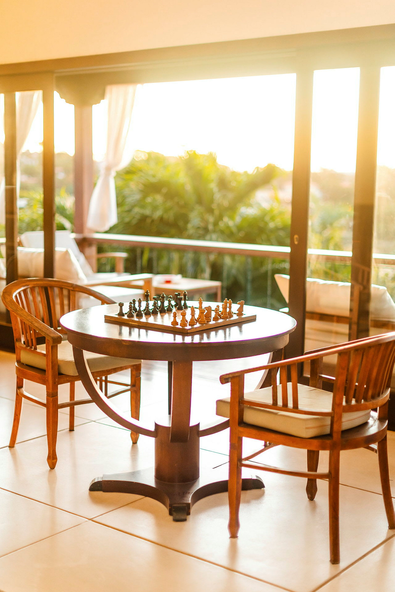 Et skakbræt står på et bord omgivet af stole på en terrasse med udsigt til grønne omgivelser. (AI-genereret tekst)