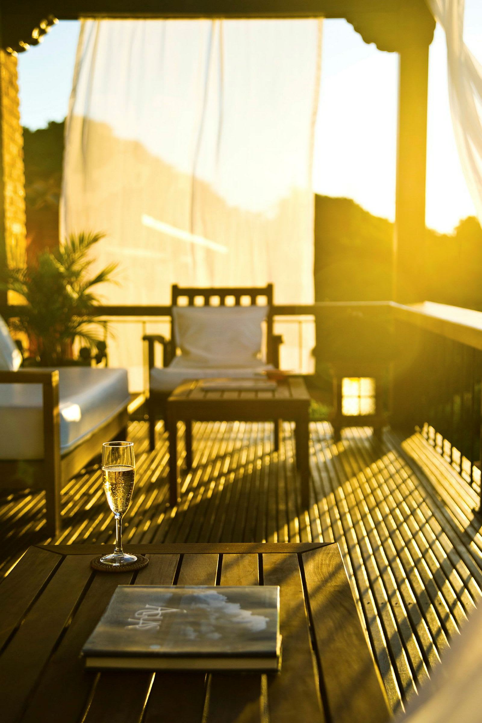 En terrasse med møbler, herunder et bord, en stol og en sofa, badet i gyldent sollys. På bordet står et glas og en bog. (AI-genereret tekst)